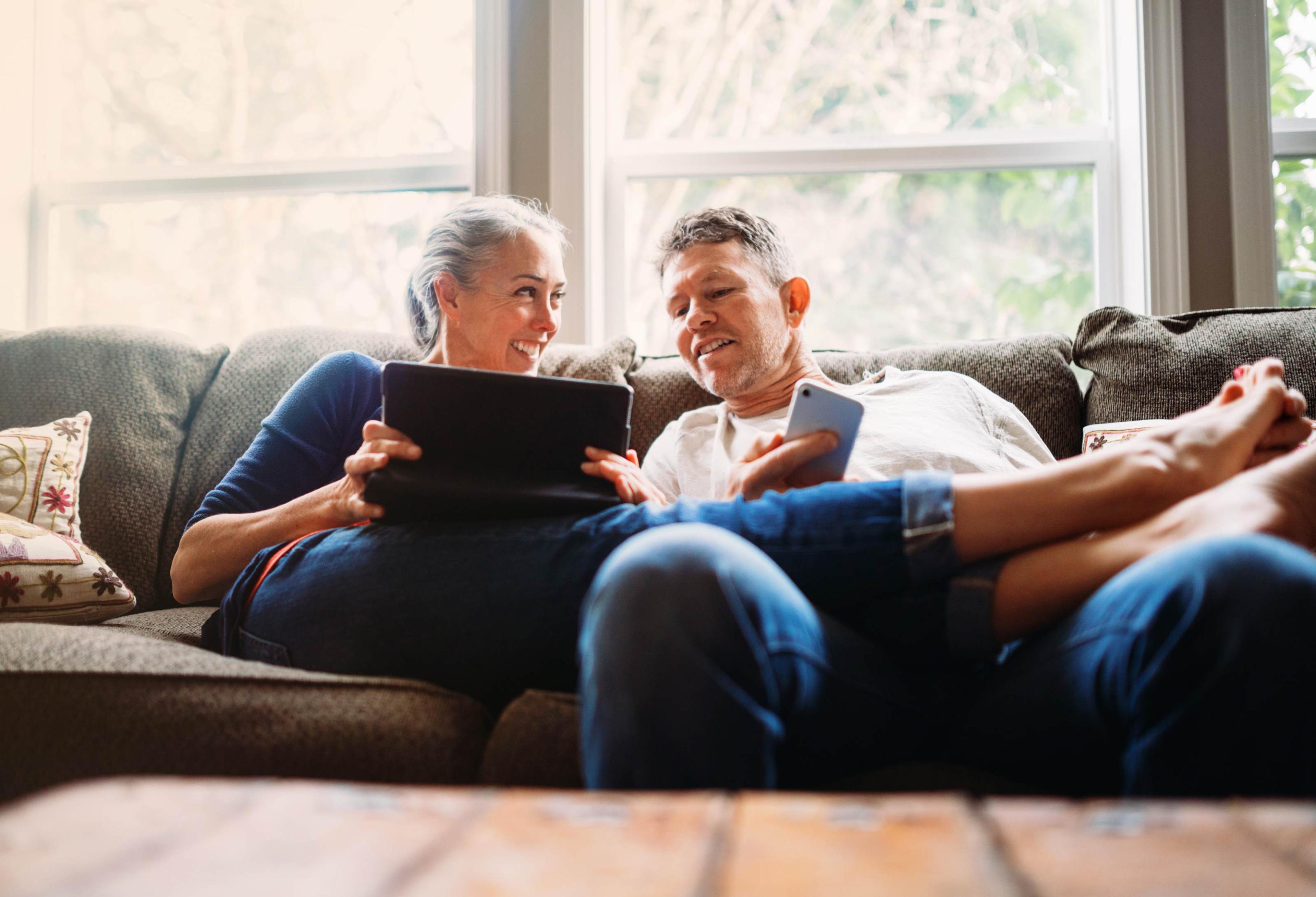Et smilende par sitter avslappet i sofaen hjemme, der kvinnen holder et nettbrett og mannen en smarttelefon mens de ser på skjermen sammen.