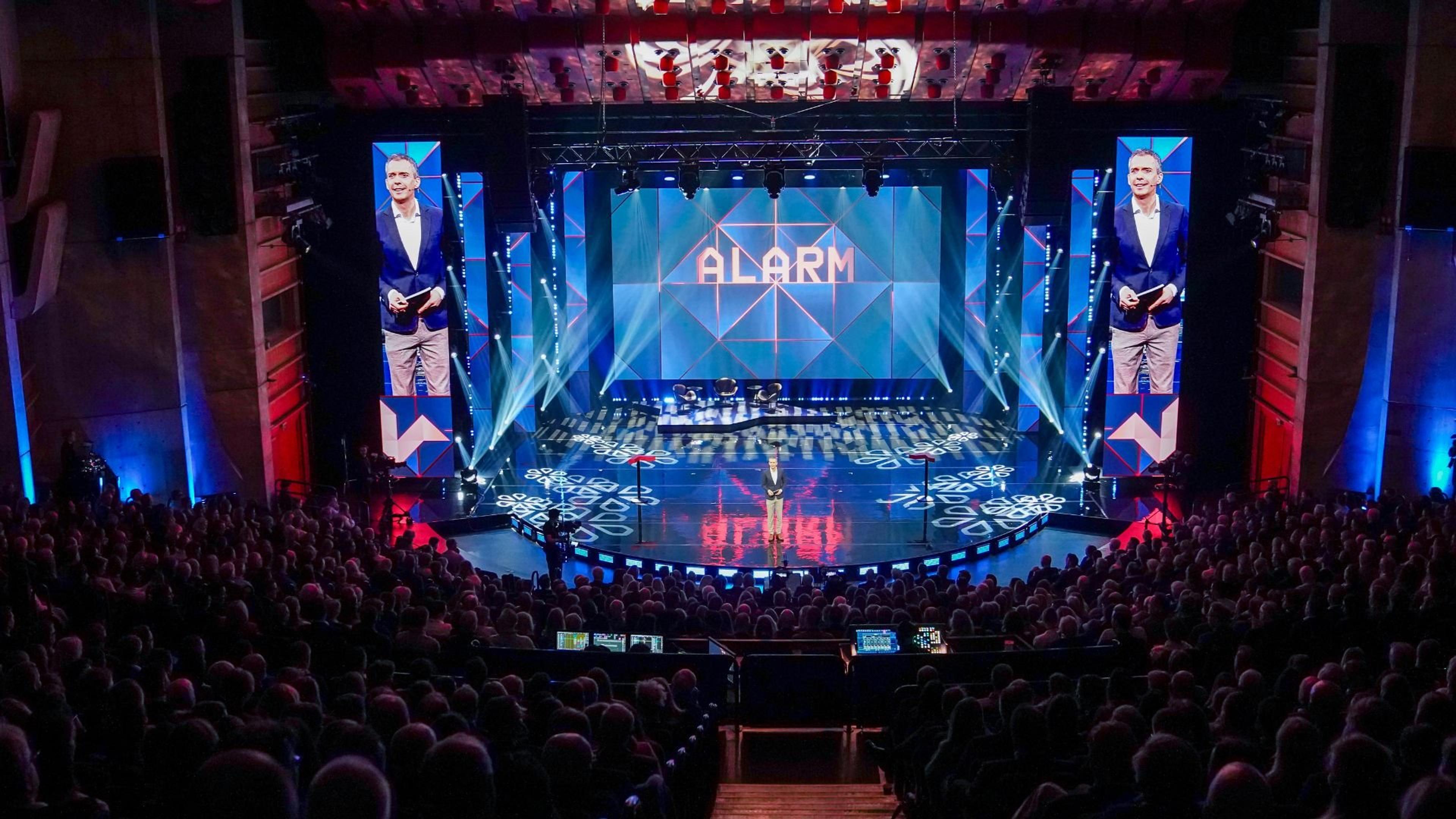 En programleder står på en opplyst scene foran et stort publikum i en sal, med ordet «ALARM» projisert på en stor skjerm bak ham og to høye skjermer som viser nærbilde av ham på hver side.