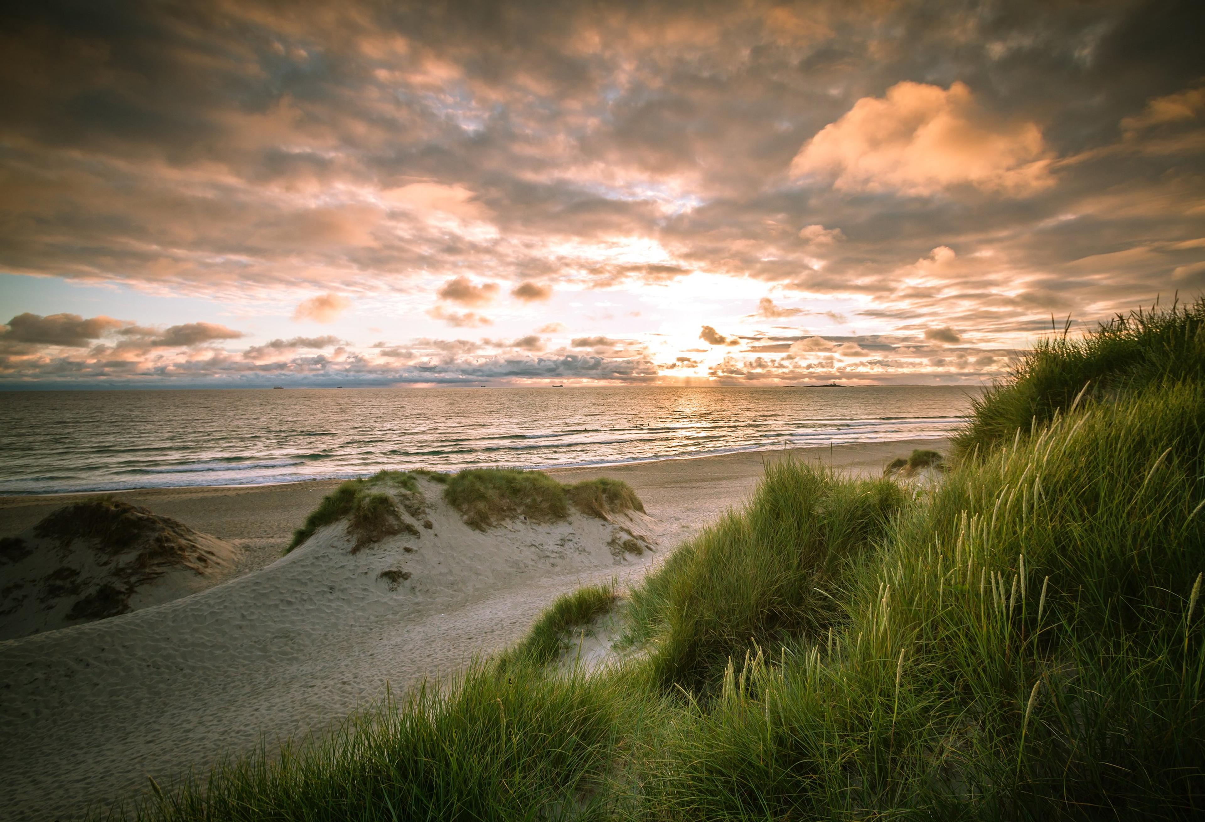 Solnedgang over havet ved en sandstrand, med gresskledde sanddyner i forgrunnen og dramatiske skyer på himmelen.