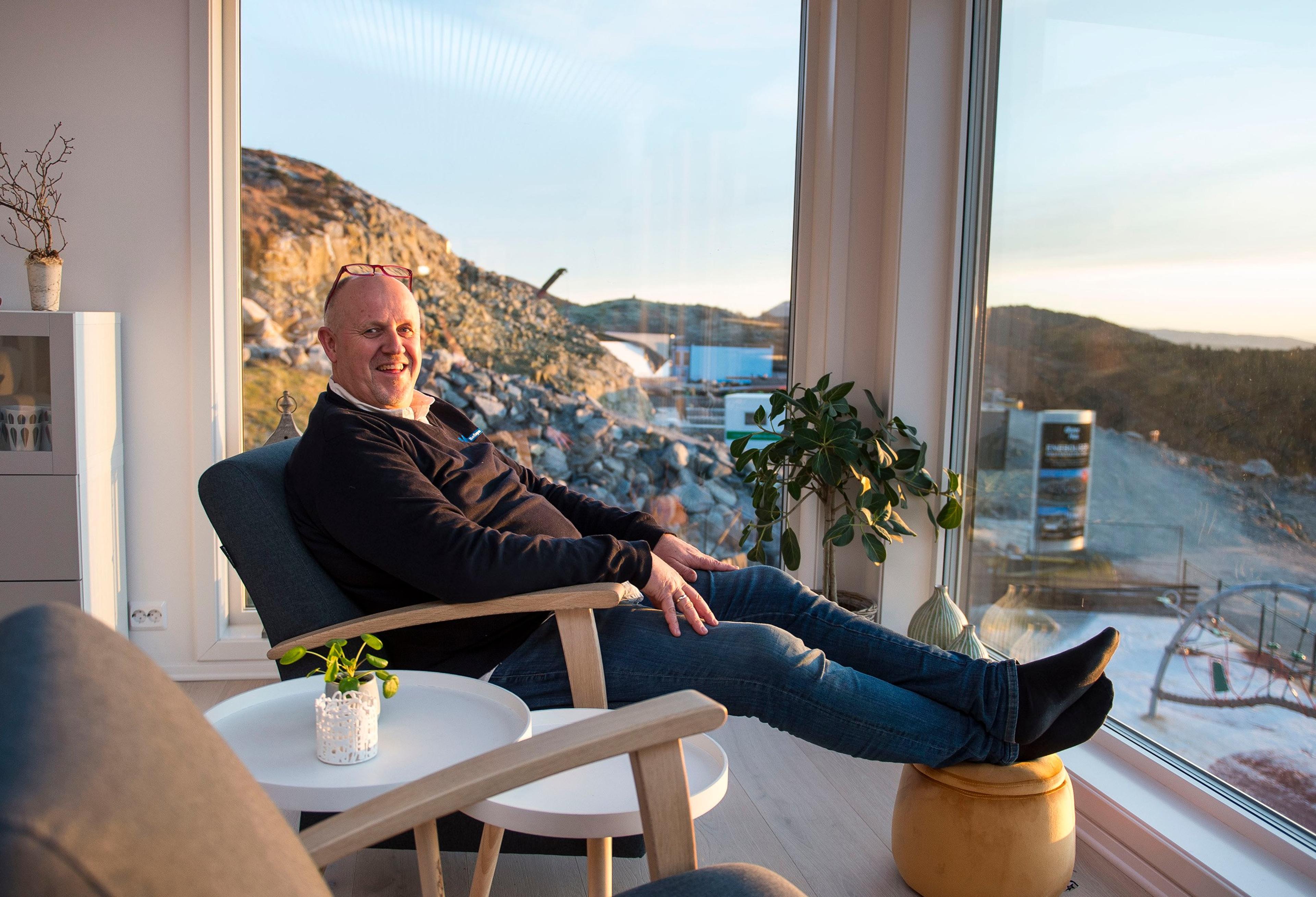 En smilende mann sitter avslappet i en lenestol med føttene på en puff foran et stort stuevindu, med utsikt mot et steinete landskap i kveldssol.