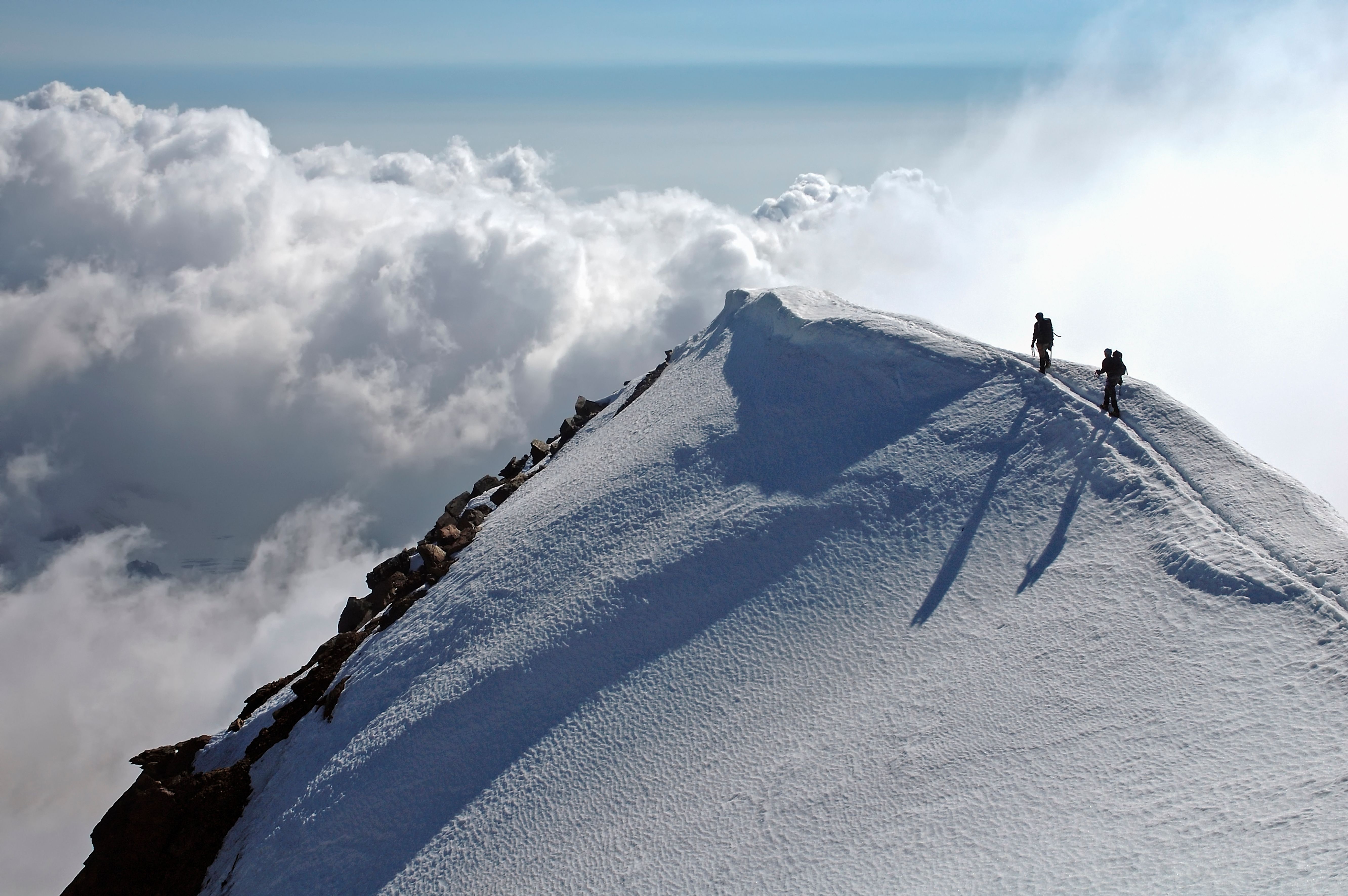 To fjellklatrere går langs en smal, snødekt fjellrygg høyt over skyene, mens lange skygger faller over snøen.