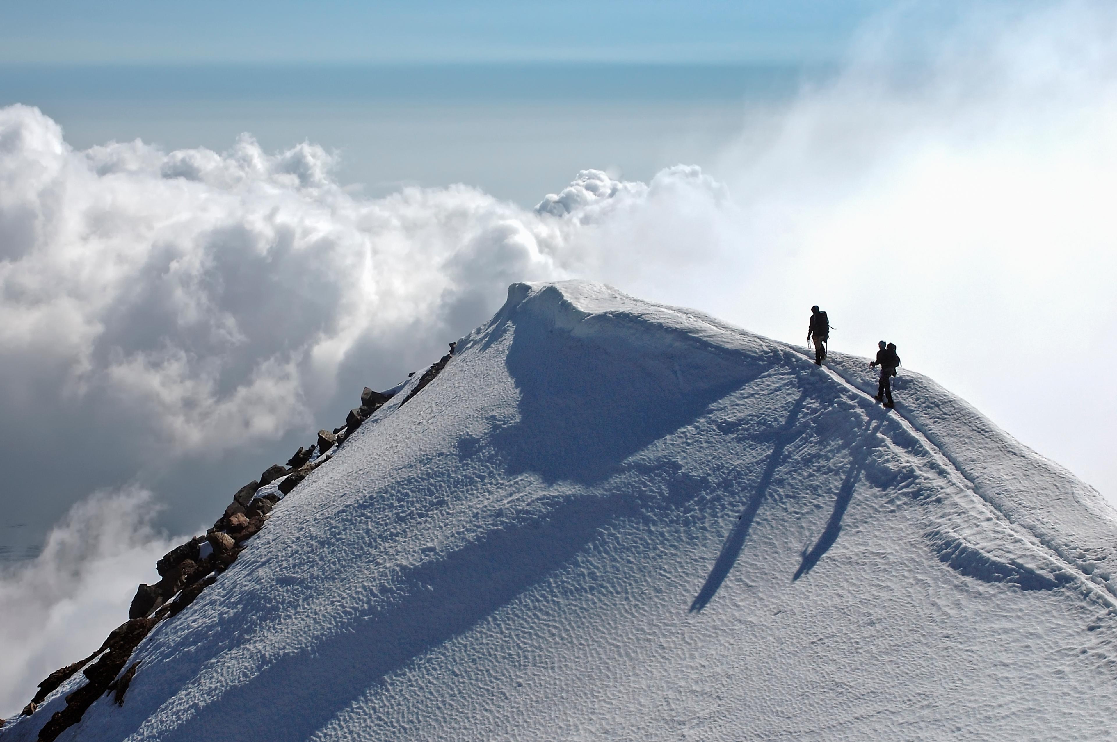 To fjellklatrere går langs en smal, snødekt fjellrygg høyt over skyene, mens lange skygger faller over snøen.