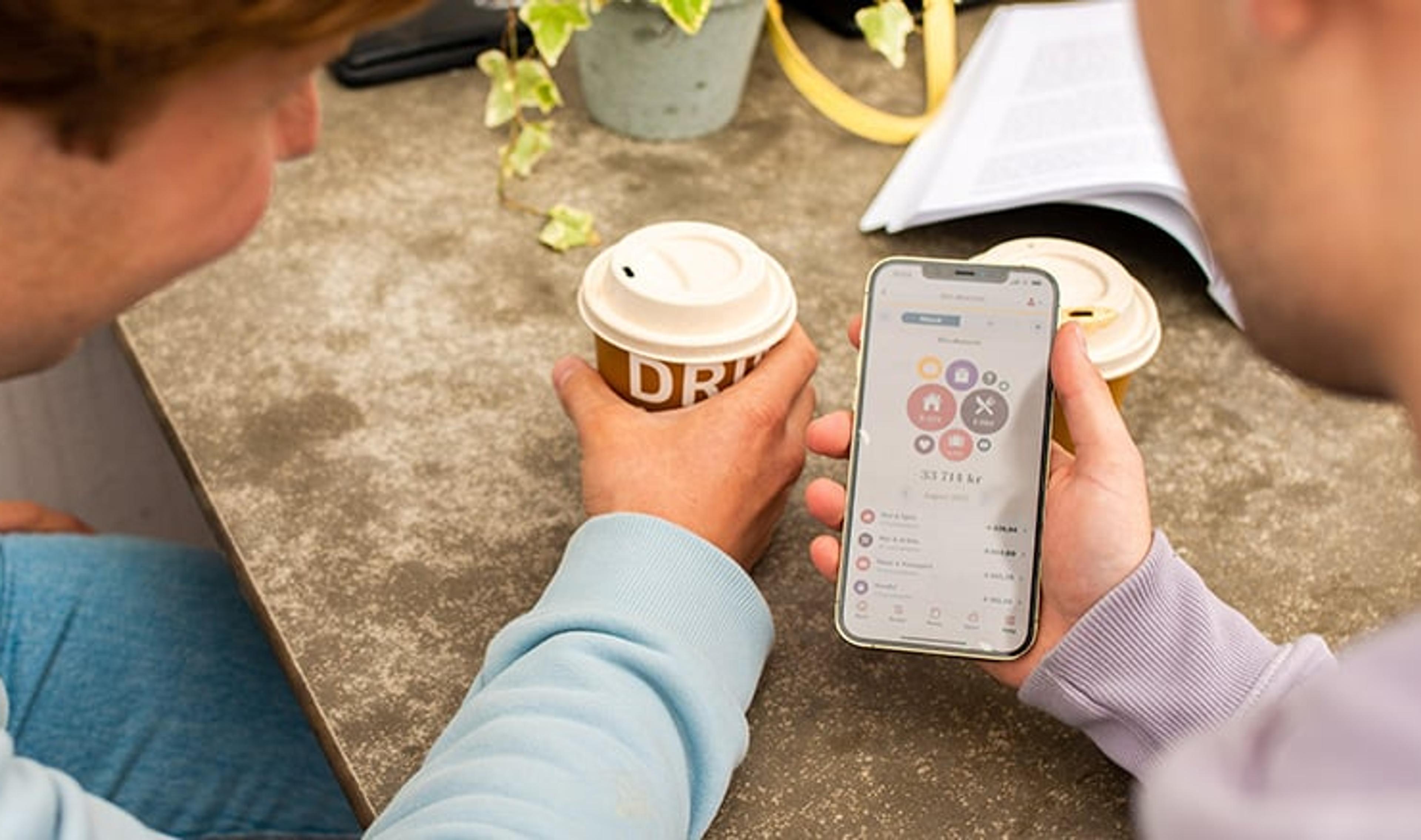 To personer sitter ved et bord med kaffekopper og ser på en mobiltelefon som viser en app med økonomisk oversikt.