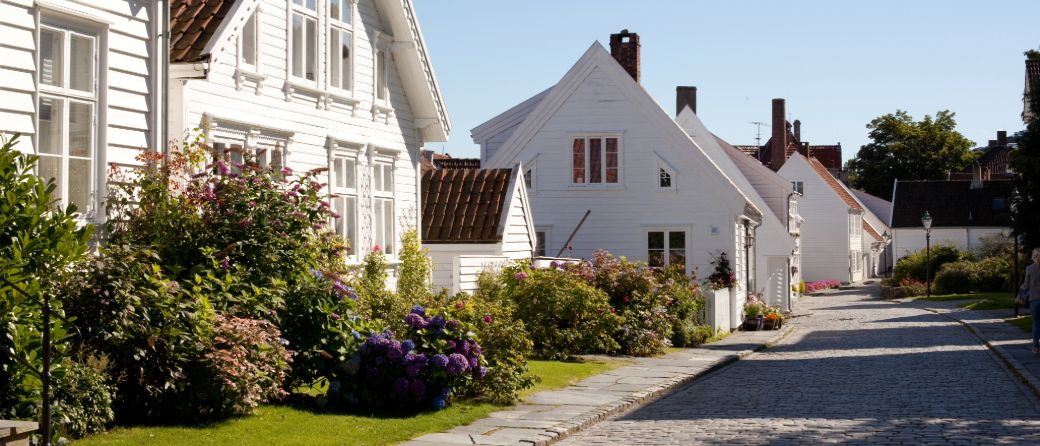 Brosteinsgate med hvitmalte trehus og frodige hager fulle av blomster i solskinn.