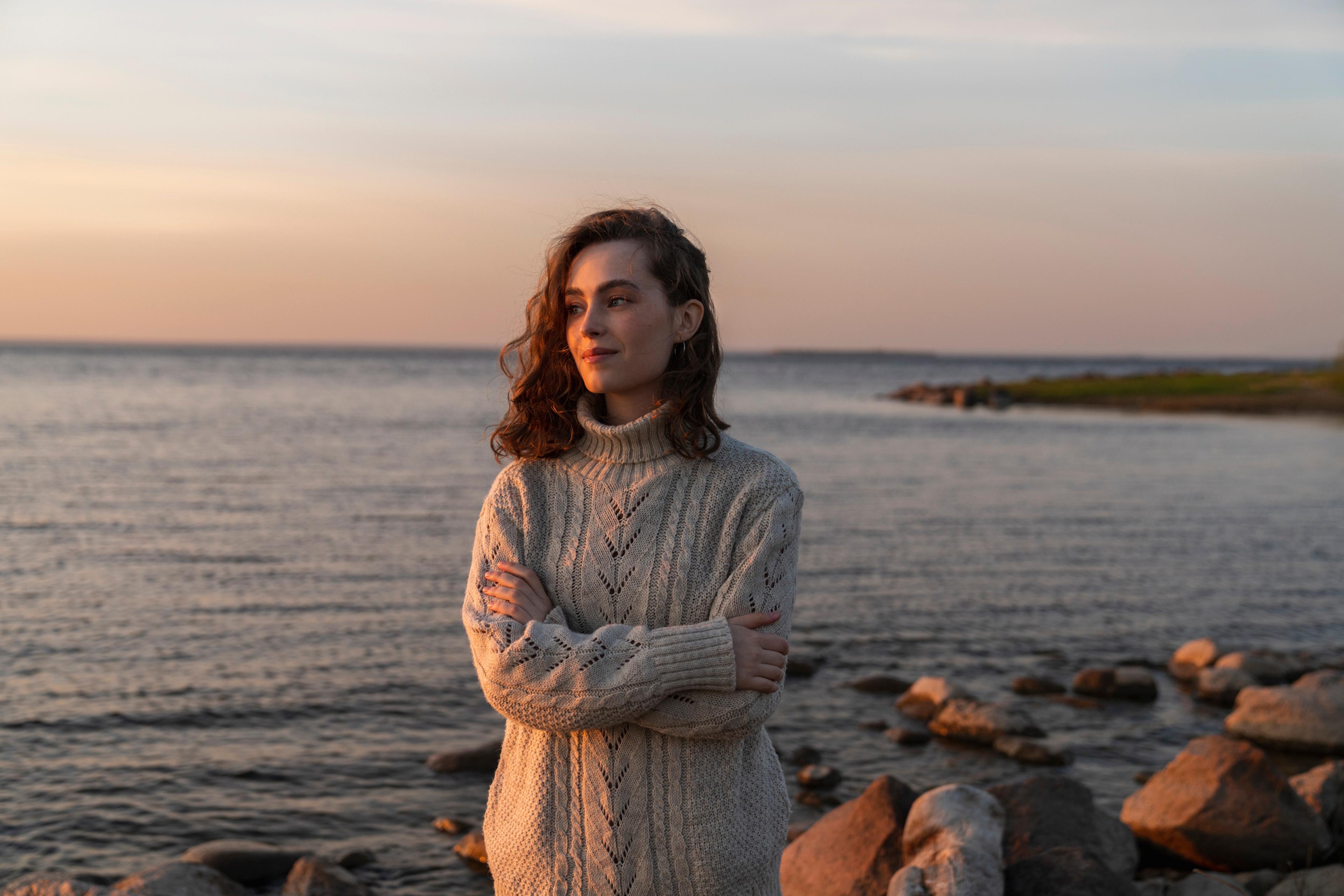 En kvinne i en lys, strikket genser står med armene i kors på en steinete strand ved solnedgang. Hun ser til siden mens varmt kveldslys faller over ansiktet og havet bak henne.