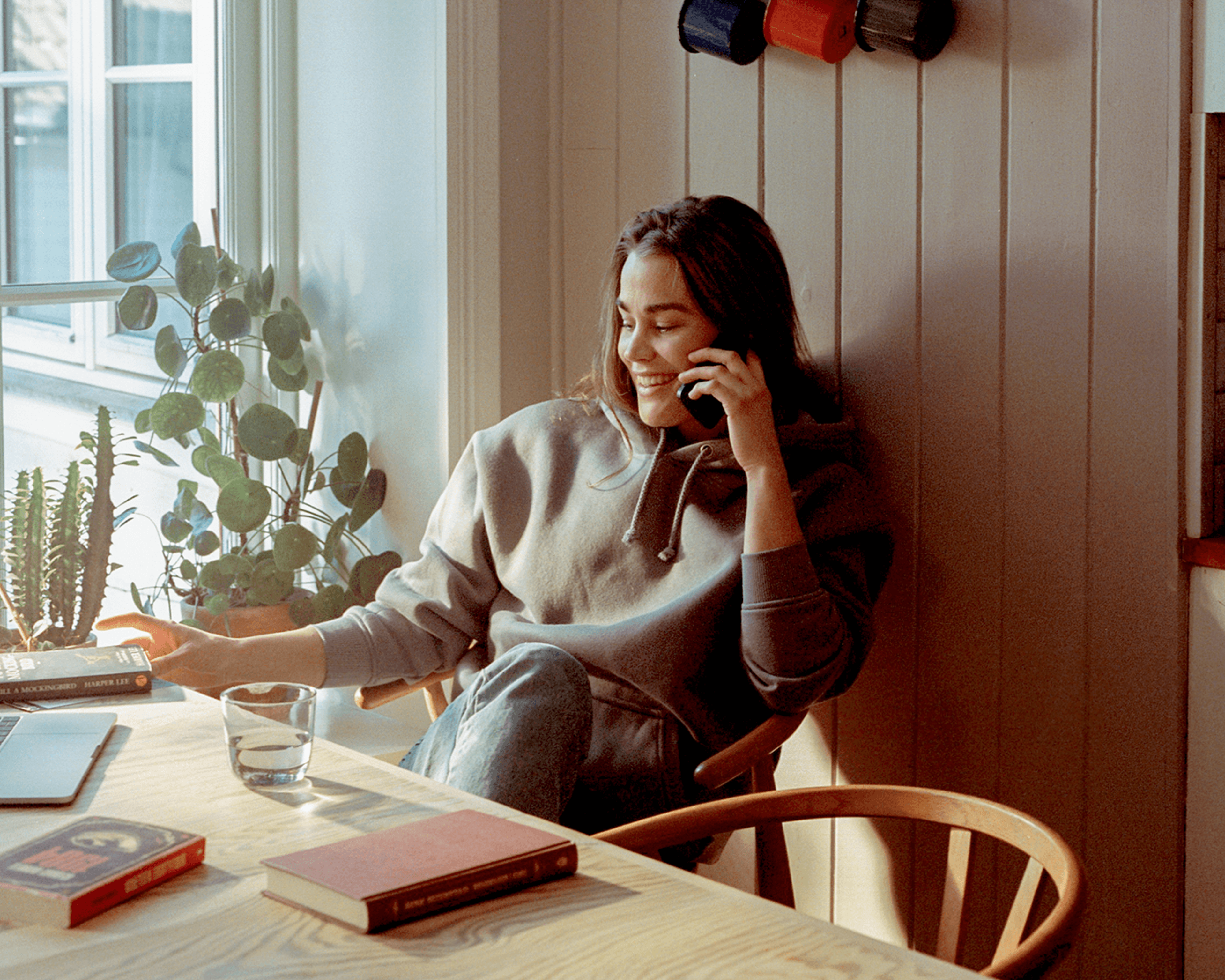 En smilende kvinne sitter ved et solfylt bord og snakker i telefonen. På bordet ligger bøker, en bærbar datamaskin og et glass vann, med grønne planter ved vinduet.
