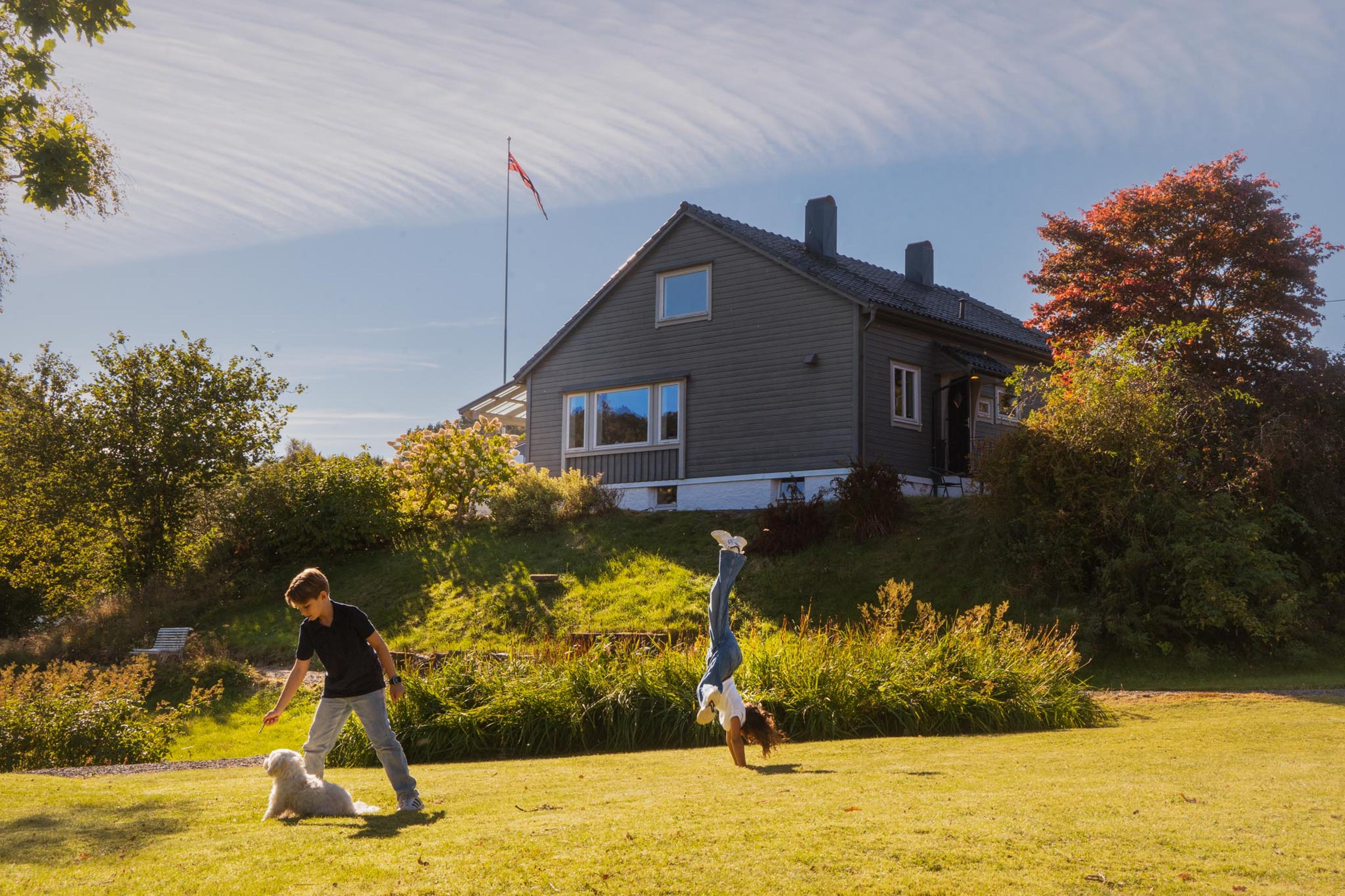 To barn leker med en liten hvit hund på plenen foran et grått hus, mens det ene barnet står på hendene. Høstfargede trær og et norsk flagg omgir huset i solskinnet.