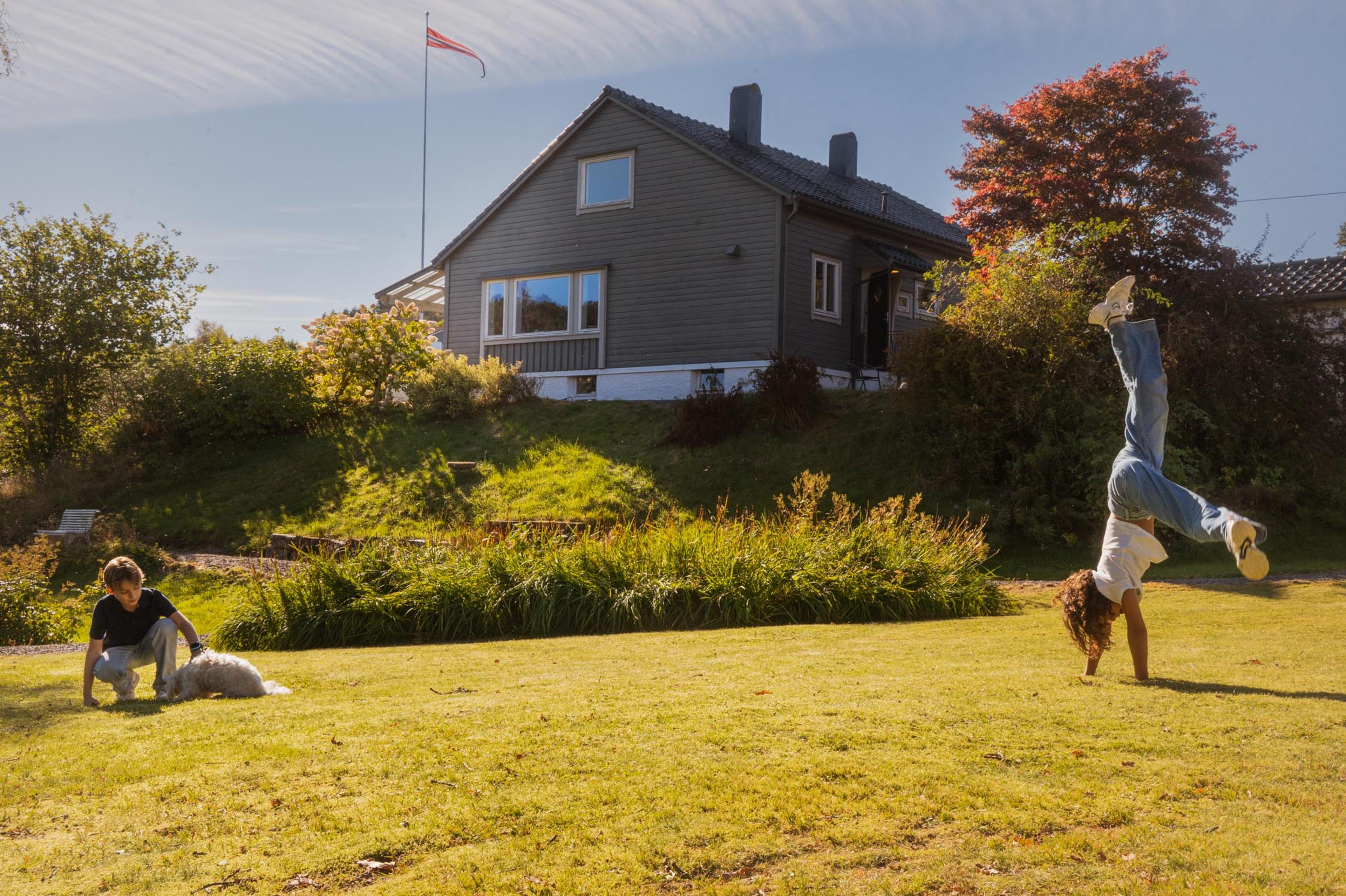 To barn leker med en liten hvit hund på en solfylt plen, mens det ene barnet står på hendene. I bakgrunnen ligger et grått hus omgitt av busker og høstfargede trær.