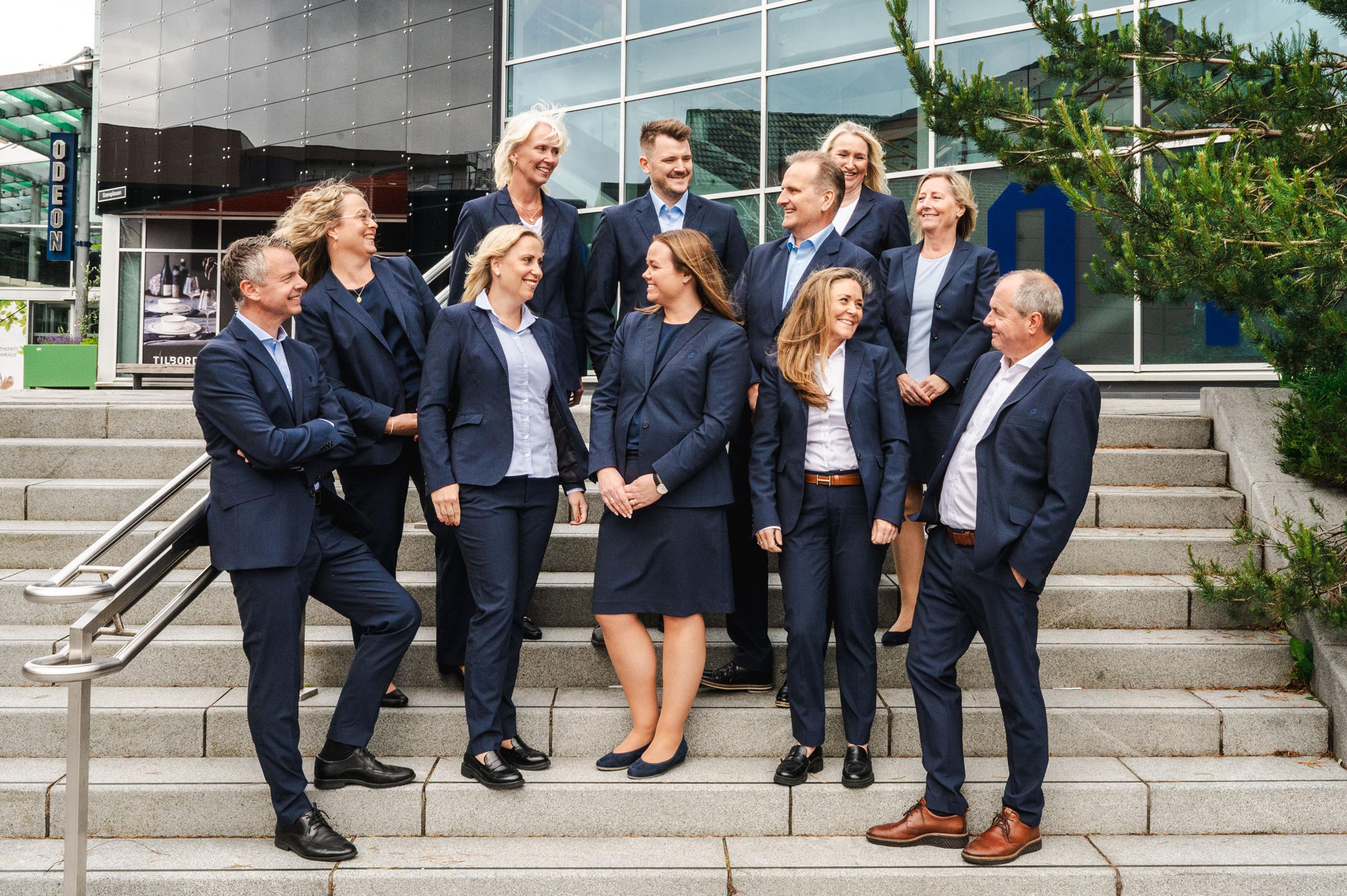 En gruppe på elleve personer i mørkeblå dresser står smilende sammen på en trapp foran en moderne glassbygning.