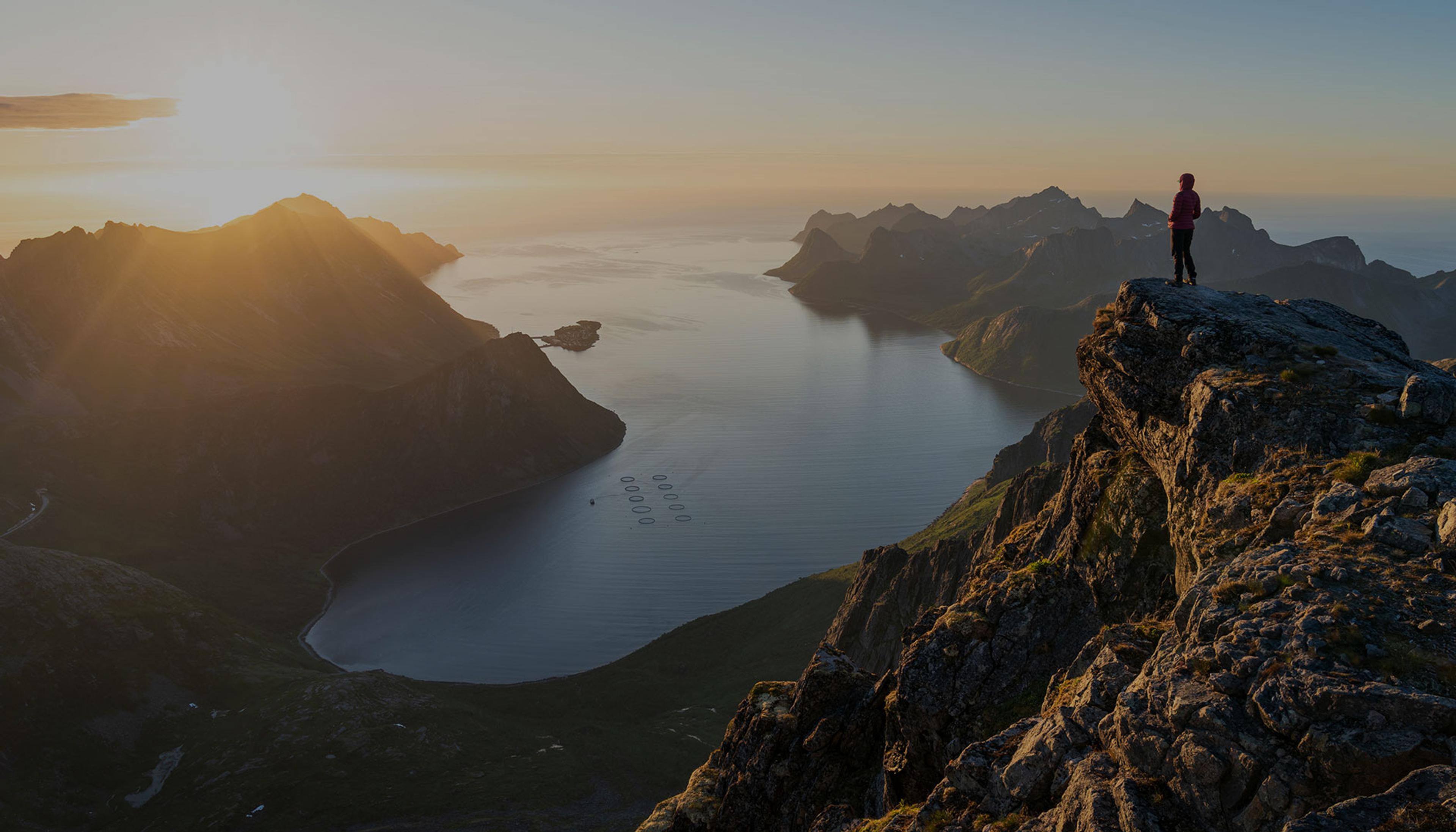 En person står på en fjellknaus og ser utover en dyp fjord omgitt av bratte fjell i solnedgang, med stille vann og oppdrettsanlegg synlig nede i fjorden.