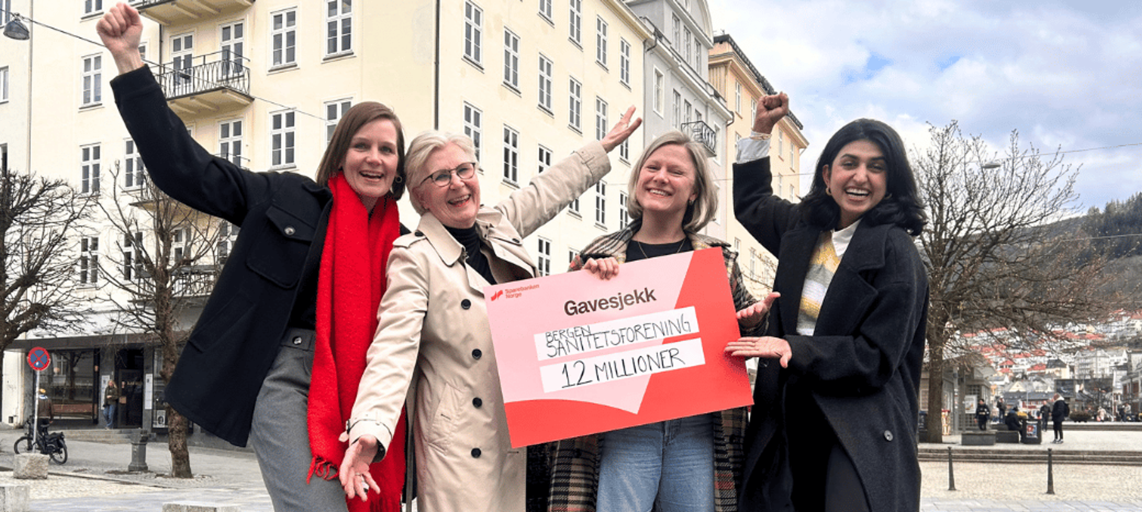 Fire jublende kvinner står på en byplass og holder en stor gavesjekk på 1,2 millioner kroner til Bergen Sanitetsforening.