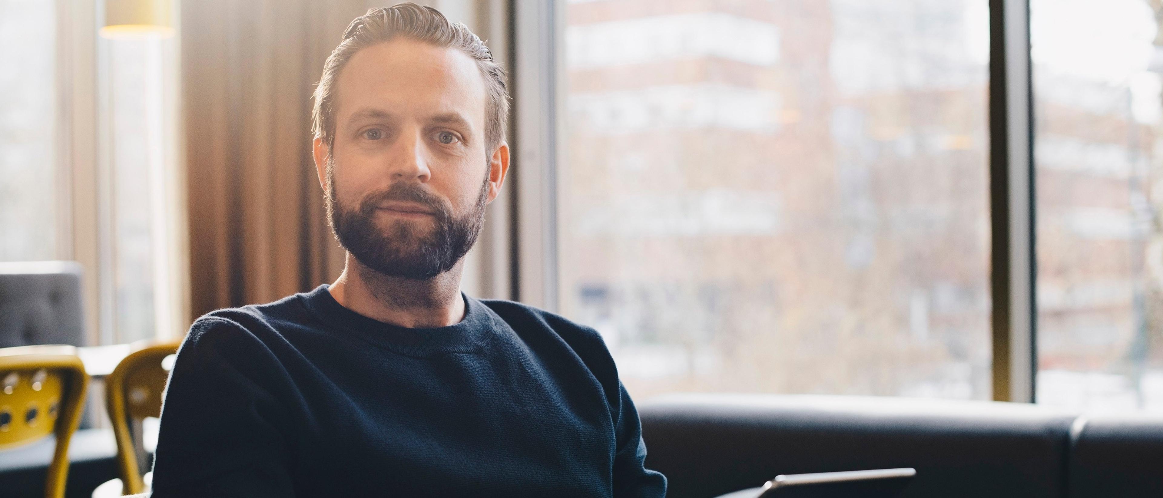 En mann med skjegg og mørk genser sitter avslappet i en sofa foran store vinduer, med et nettbrett eller en bærbar PC foran seg i et lyst kontor- eller kafémiljø.