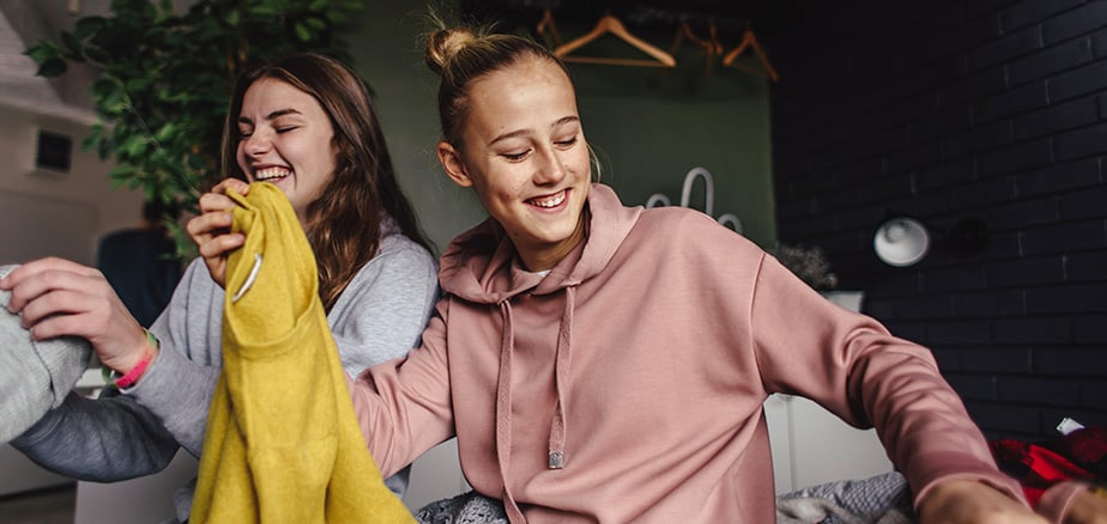 To smilende unge kvinner sitter sammen i et rom; den ene holder opp et gult klesplagg, mens den andre har på seg en rosa hettegenser.