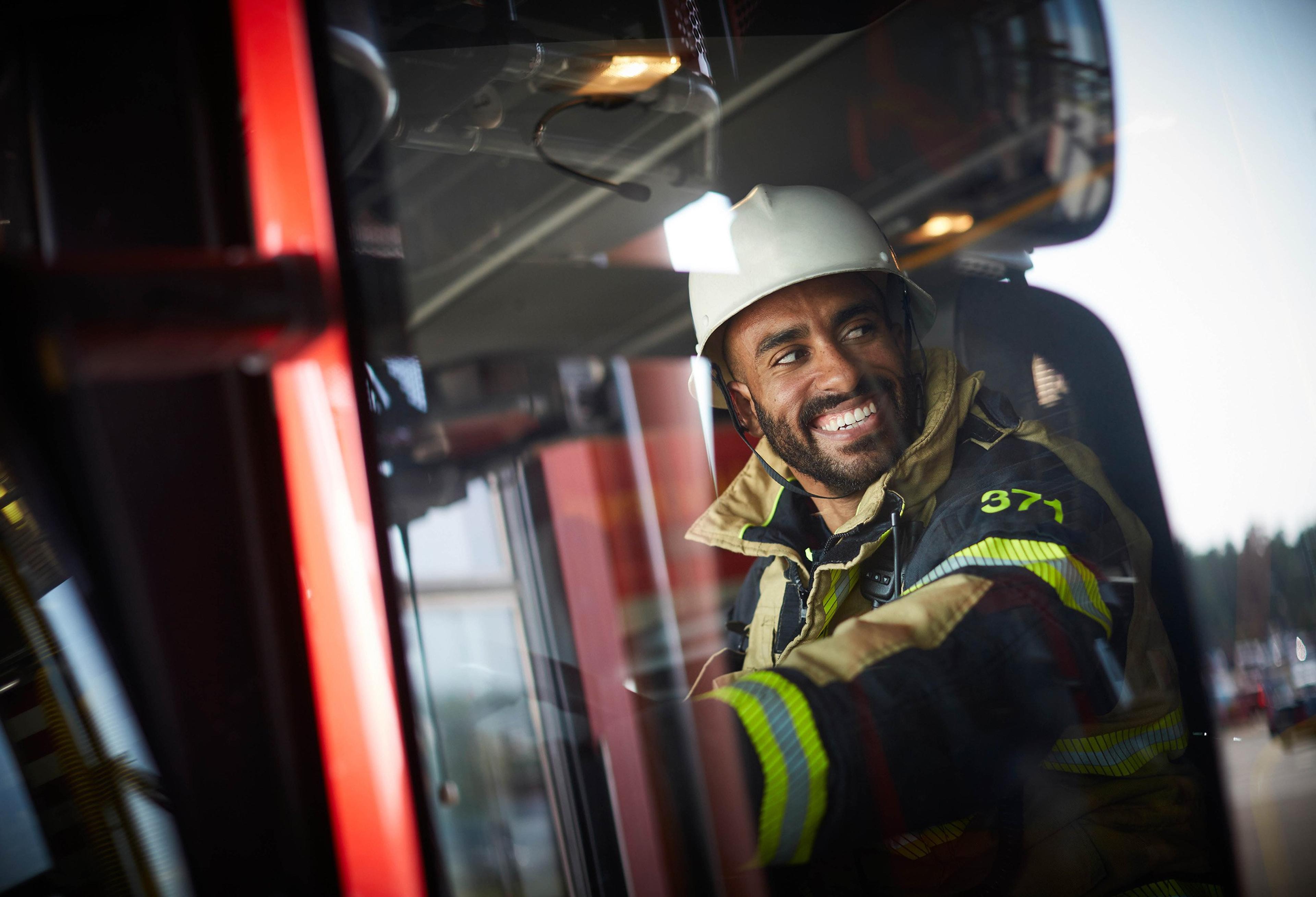 En smilende brannmann med hvit hjelm og verneklær sitter i førerhuset på en rød brannbil.