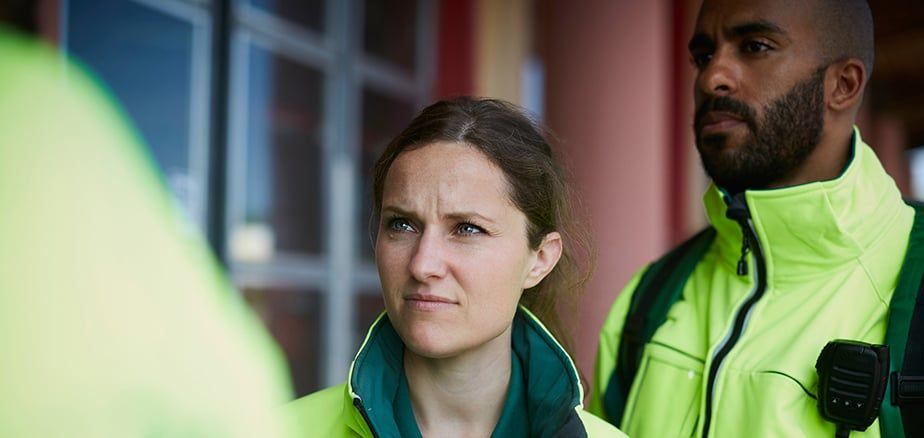 To redningsarbeidere i neongule uniformer lytter alvorlig til en person utenfor bildet. Mannen til høyre har en radiosender festet til uniformen.