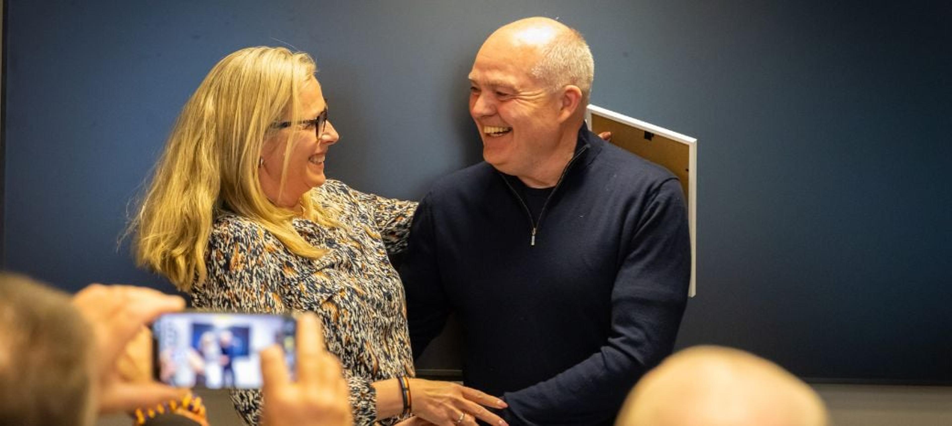 En smilende kvinne og mann står med armene rundt hverandre foran en mørk vegg, mens noen fotograferer dem med en mobiltelefon.