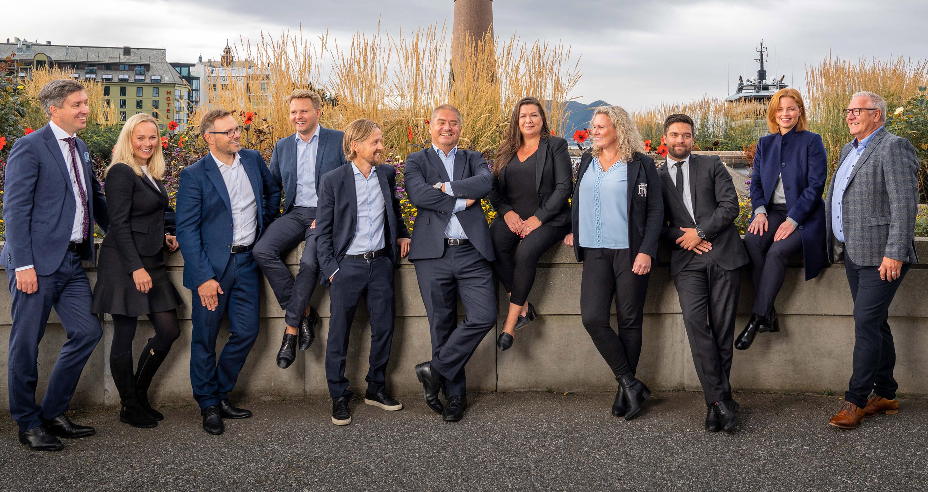 En gruppe på elleve forretningsfolk står og sitter avslappet langs en mur foran blomster og høyt gress, kledd i formelle klær og smilende mot kameraet i et urbant utendørsmiljø.