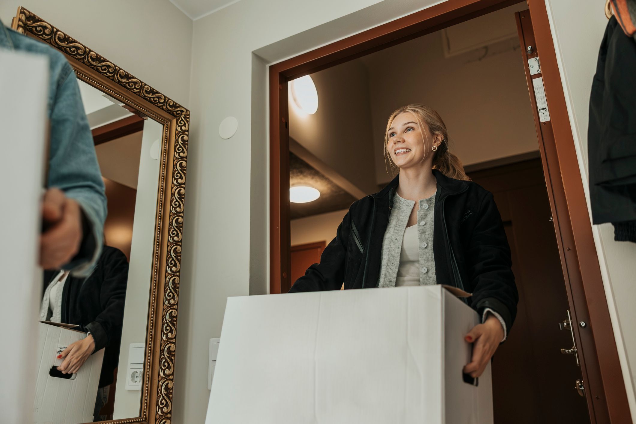 En smilende ung kvinne bærer en stor hvit pappeske inn gjennom en åpen dør. Et stort speil med dekorativ ramme står i gangen ved siden av henne.