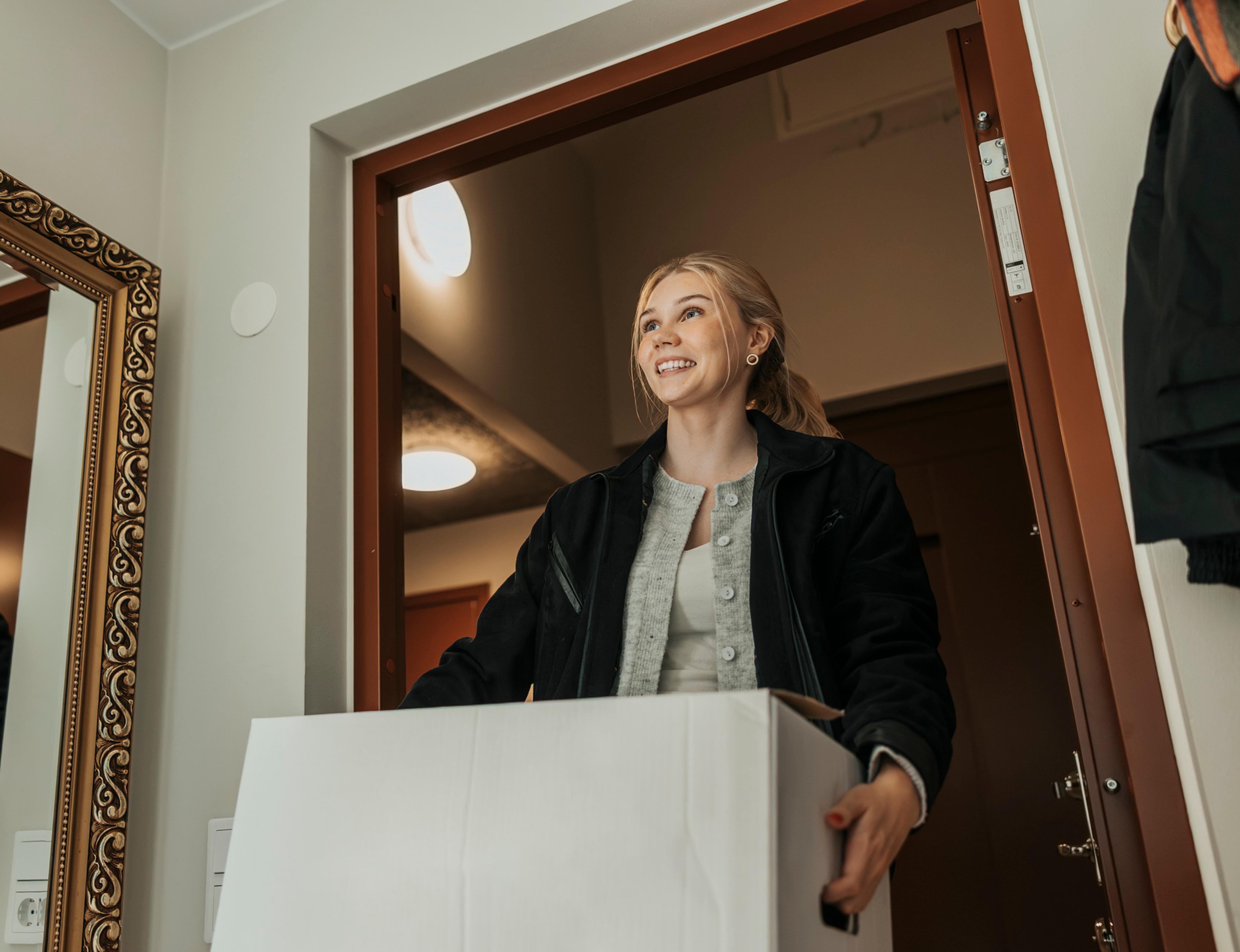 En smilende ung kvinne bærer en stor hvit pappeske inn gjennom en åpen dør. Et stort speil med dekorativ ramme står i gangen ved siden av henne.