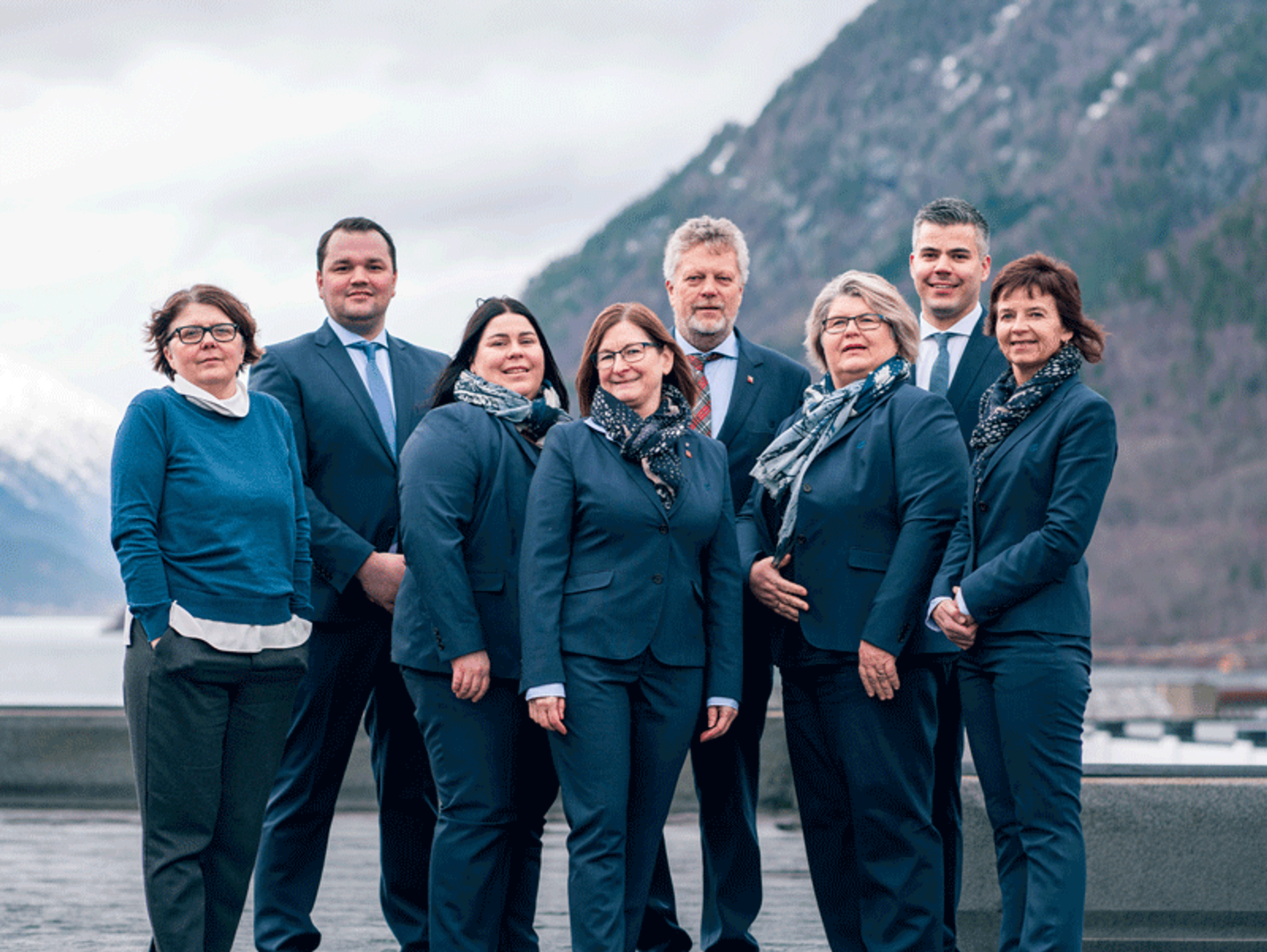 Åtte personer i formelle, mørke antrekk står samlet utendørs foran et fjellandskap.