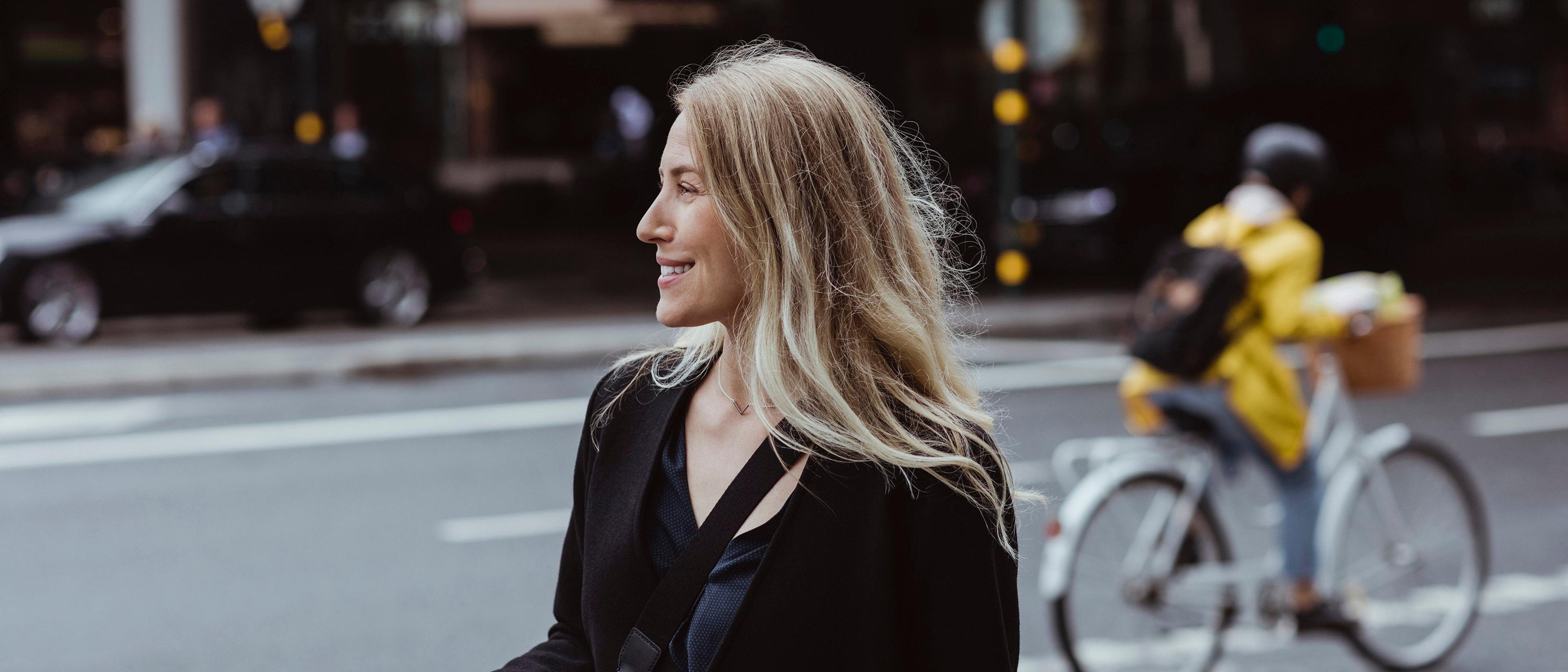 Smilende kvinne med langt blondt hår står ved en bygate og ser til siden, mens en syklist i gul jakke passerer i bakgrunnen.