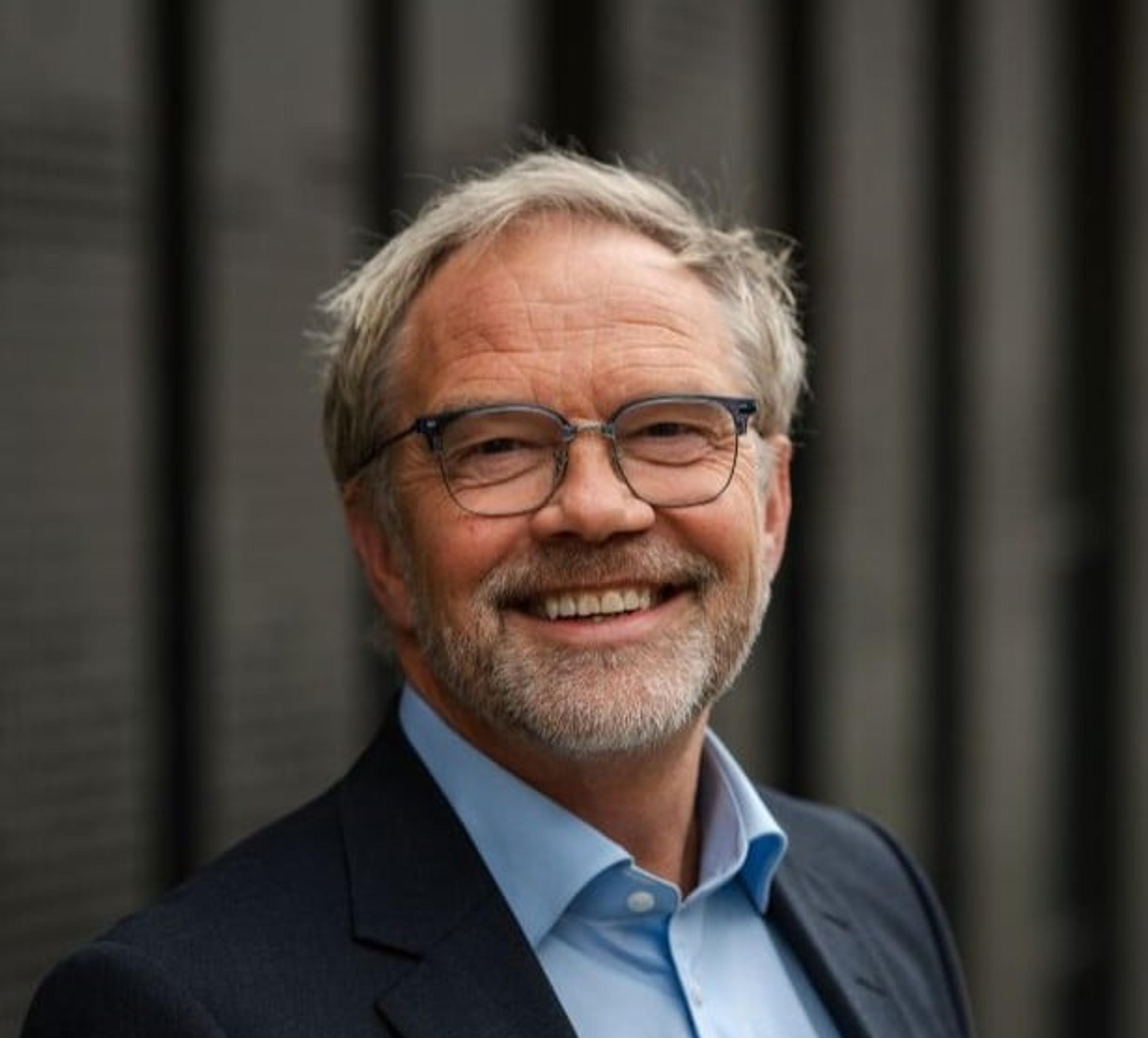 Smilende middelaldrende mann med briller, skjegg og dressjakke står foran en mørk, uskarp bakgrunn. Han ser rett mot kamera i et formelt, men vennlig portrett.
