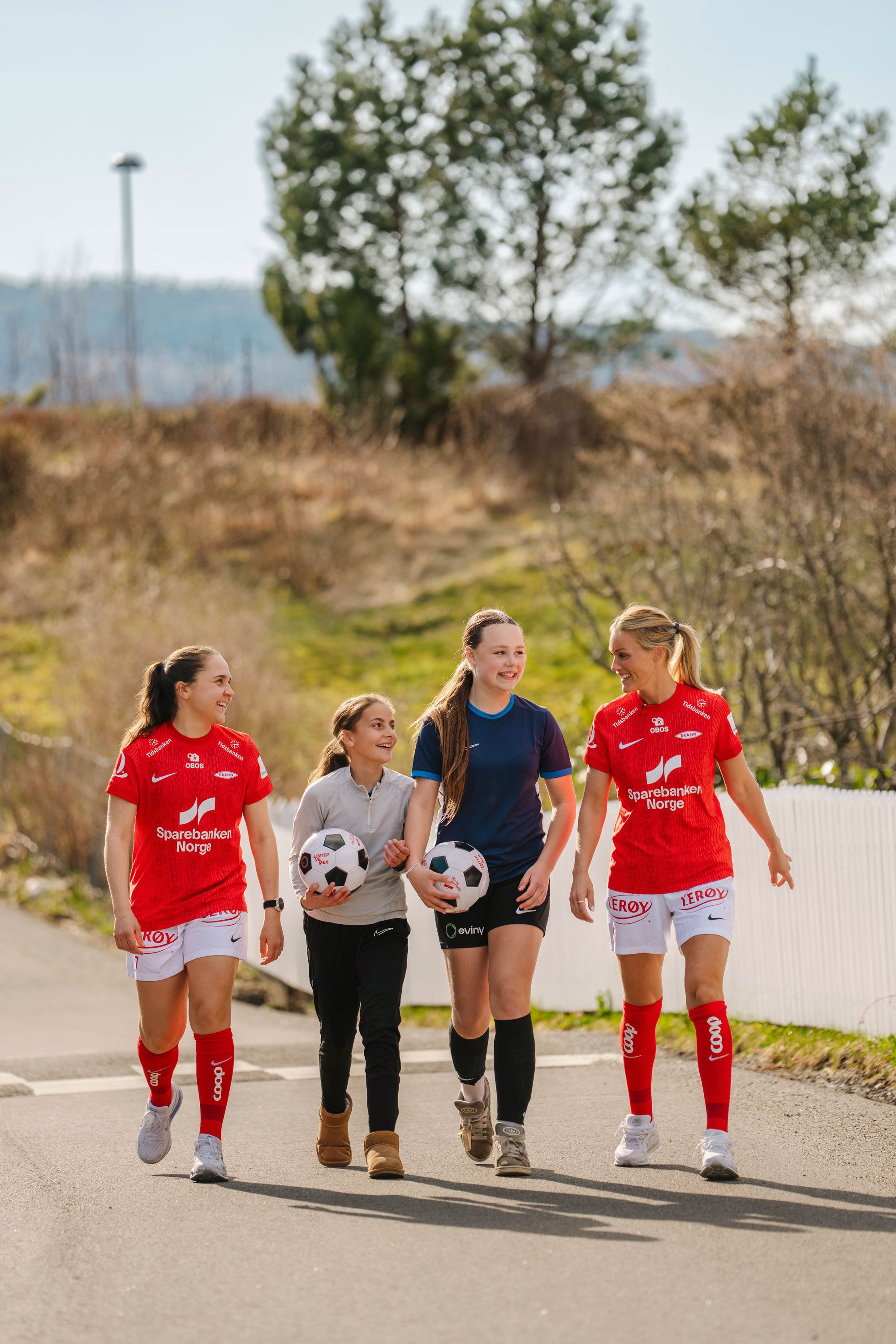 To kvinnelige fotballspillere i røde drakter går langs en vei sammen med to jenter som holder hver sin fotball. De smiler og snakker sammen i solskinnet.