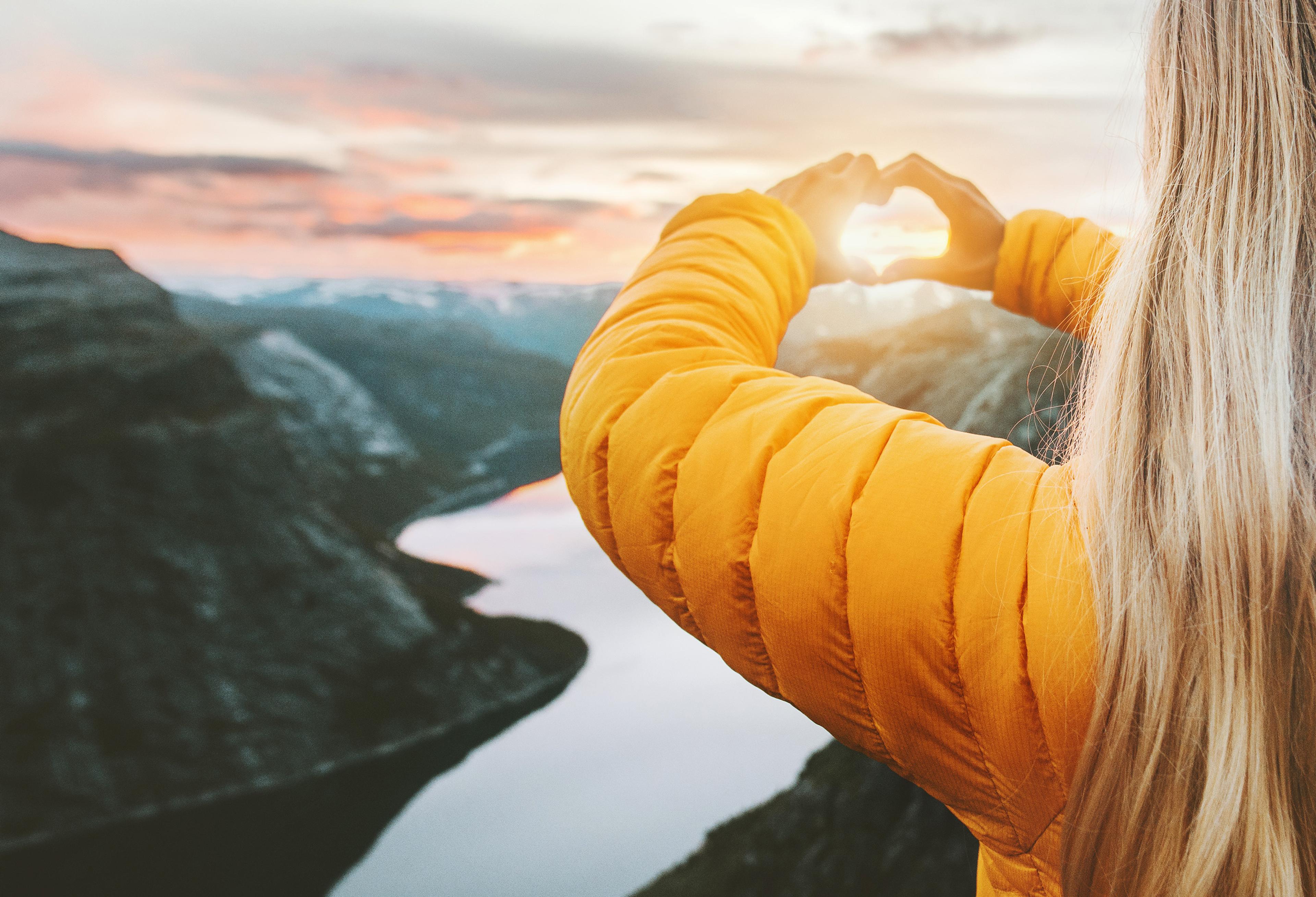 Person i gul jakke står med ryggen til kameraet og former et hjerte med hendene rundt sola over en fjord omgitt av fjell i solnedgang.