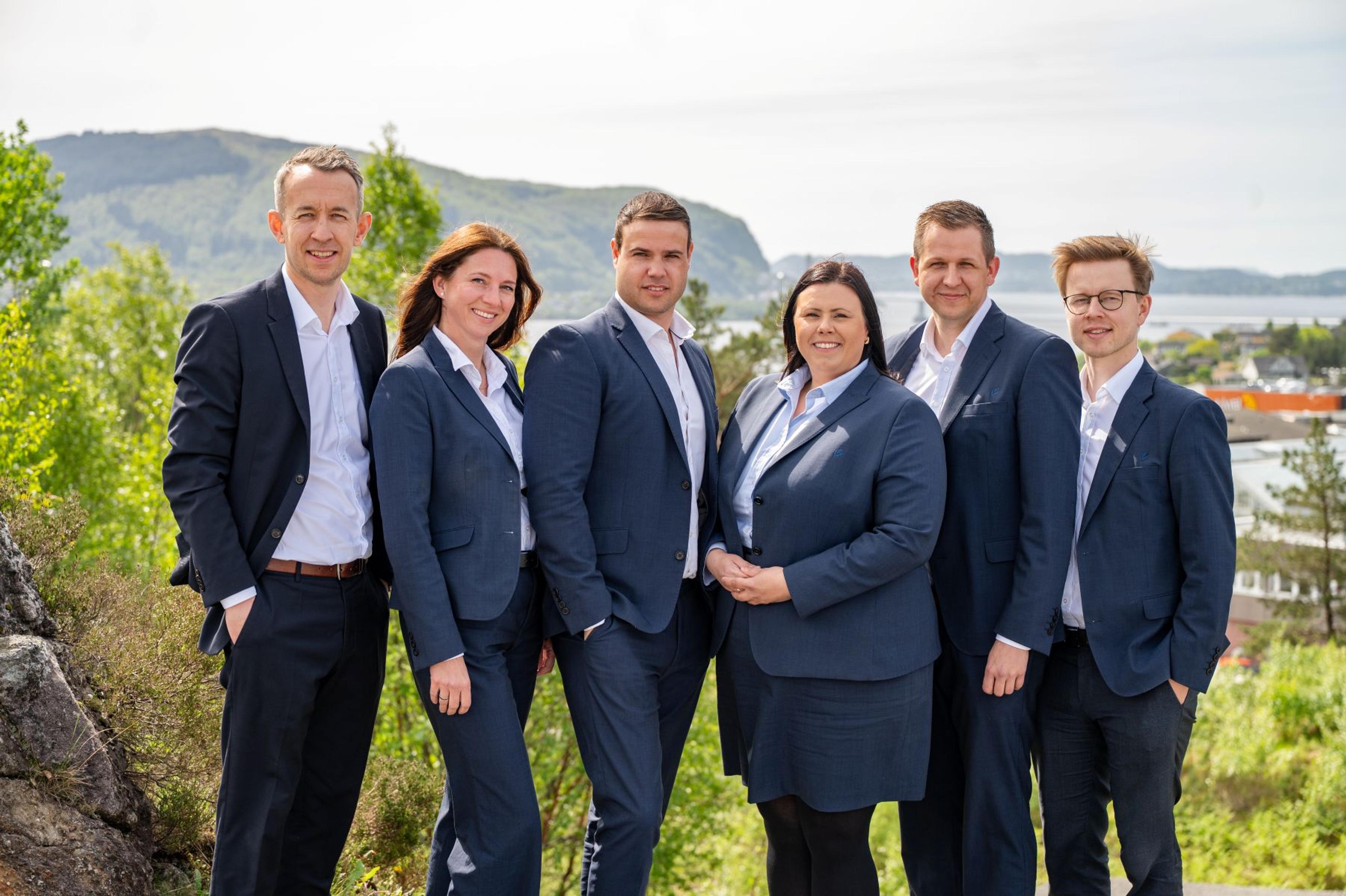 Syv personer i formelle, mørkeblå dresser står side om side utendørs og smiler mot kameraet, med grønn natur, fjell og vann i bakgrunnen.