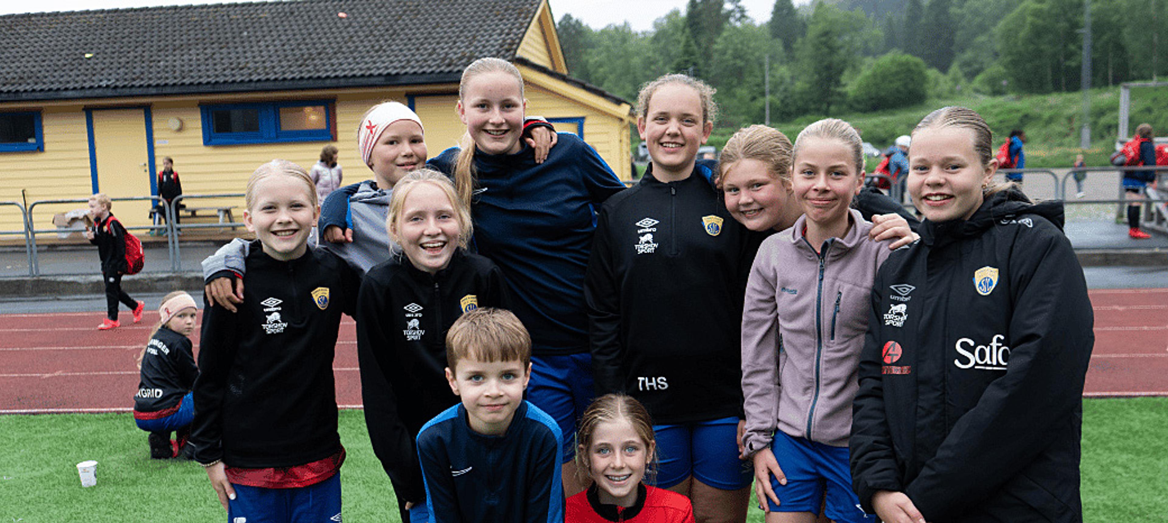 En gruppe smilende barn og ungdommer i idrettsklær står samlet på en friidrettsbane.