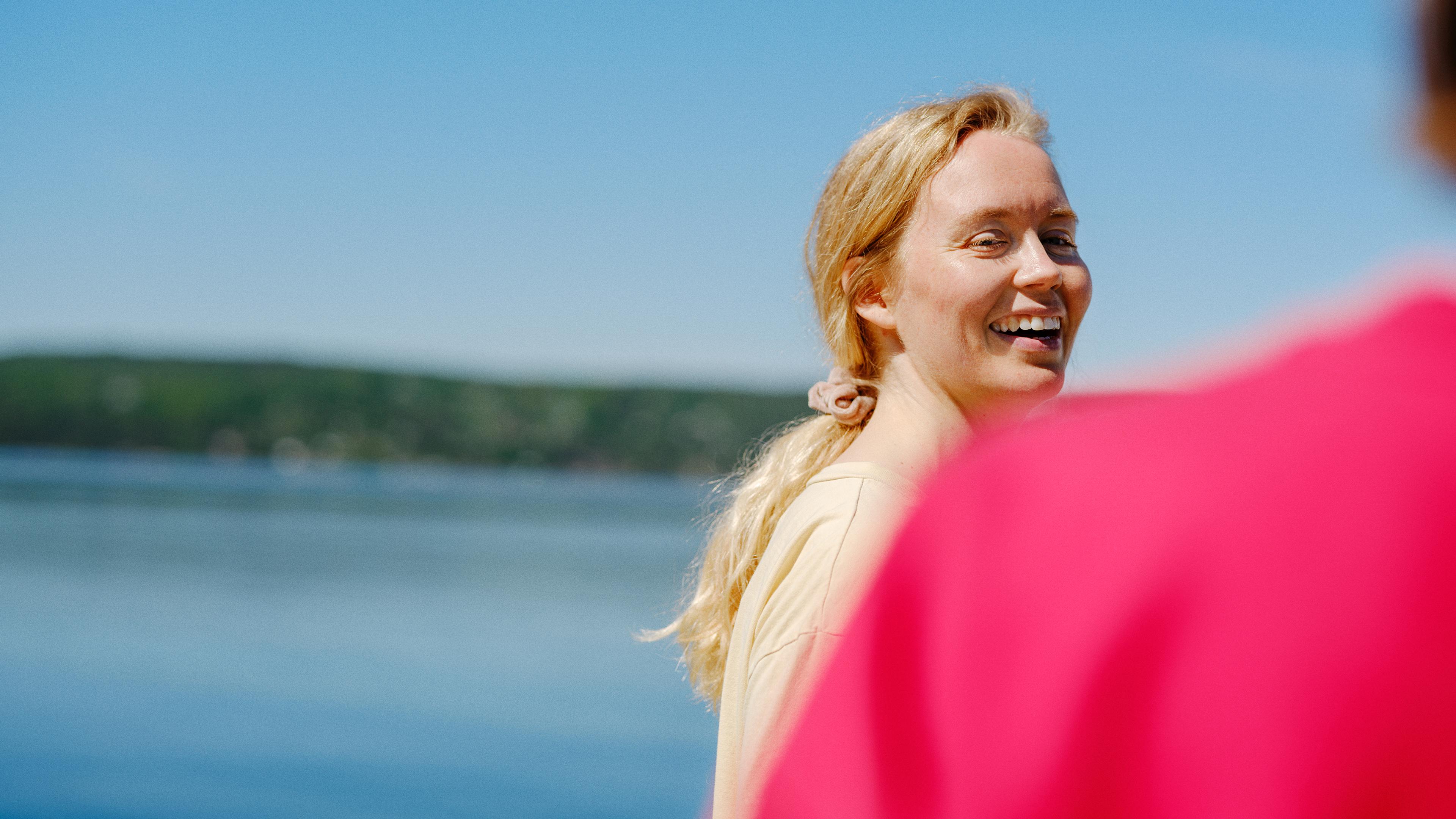 En smilende kvinne med blond hestehale står utendørs ved vannet på en solrik dag, med en person i rosa genser uskarpt i forgrunnen.