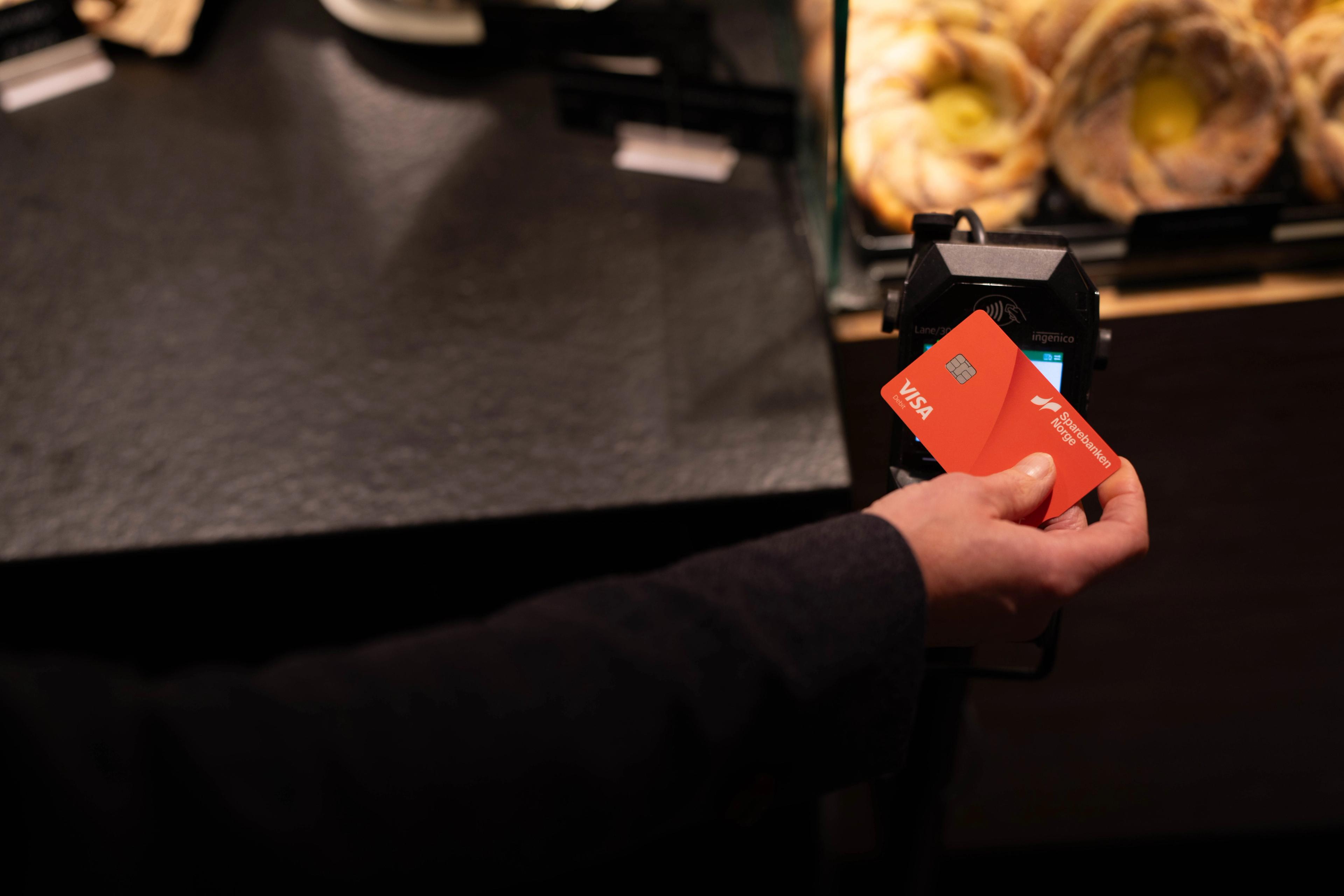 Hånd som holder en smarttelefon foran en betalingsterminal.