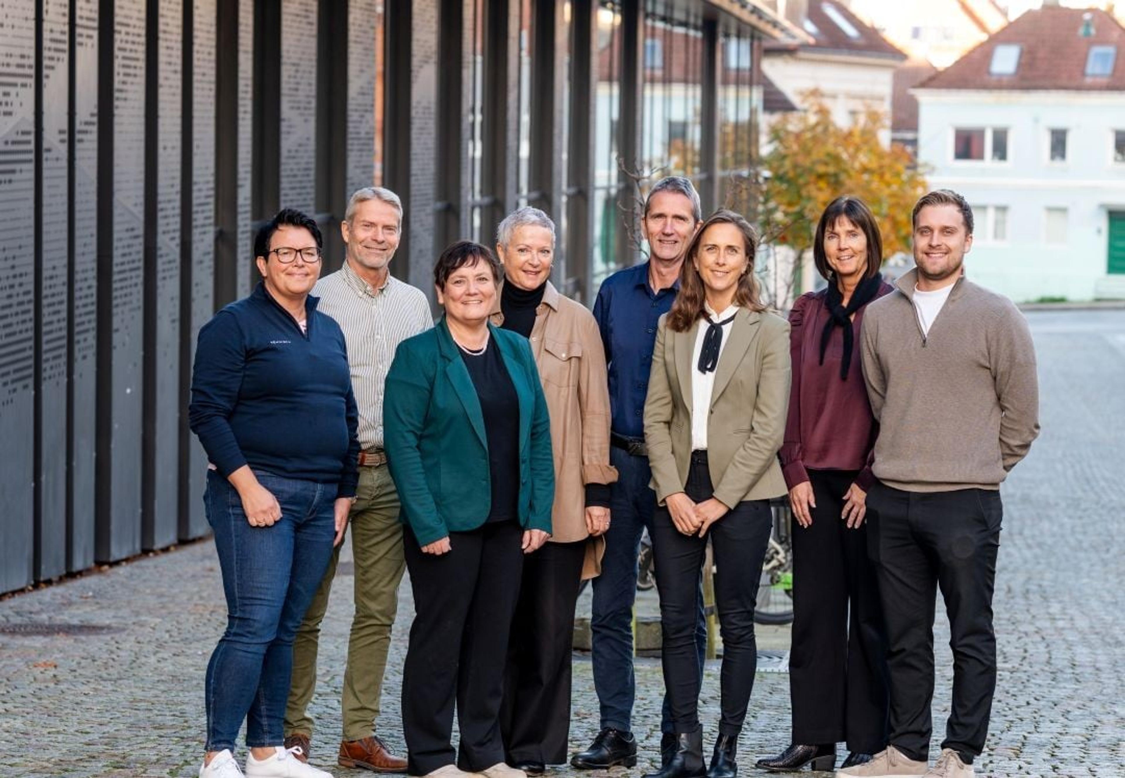 En gruppe på åtte voksne personer står ute på en brosteinsgate foran en moderne bygning, smilende mot kameraet som om de er kolleger eller et team på jobb.