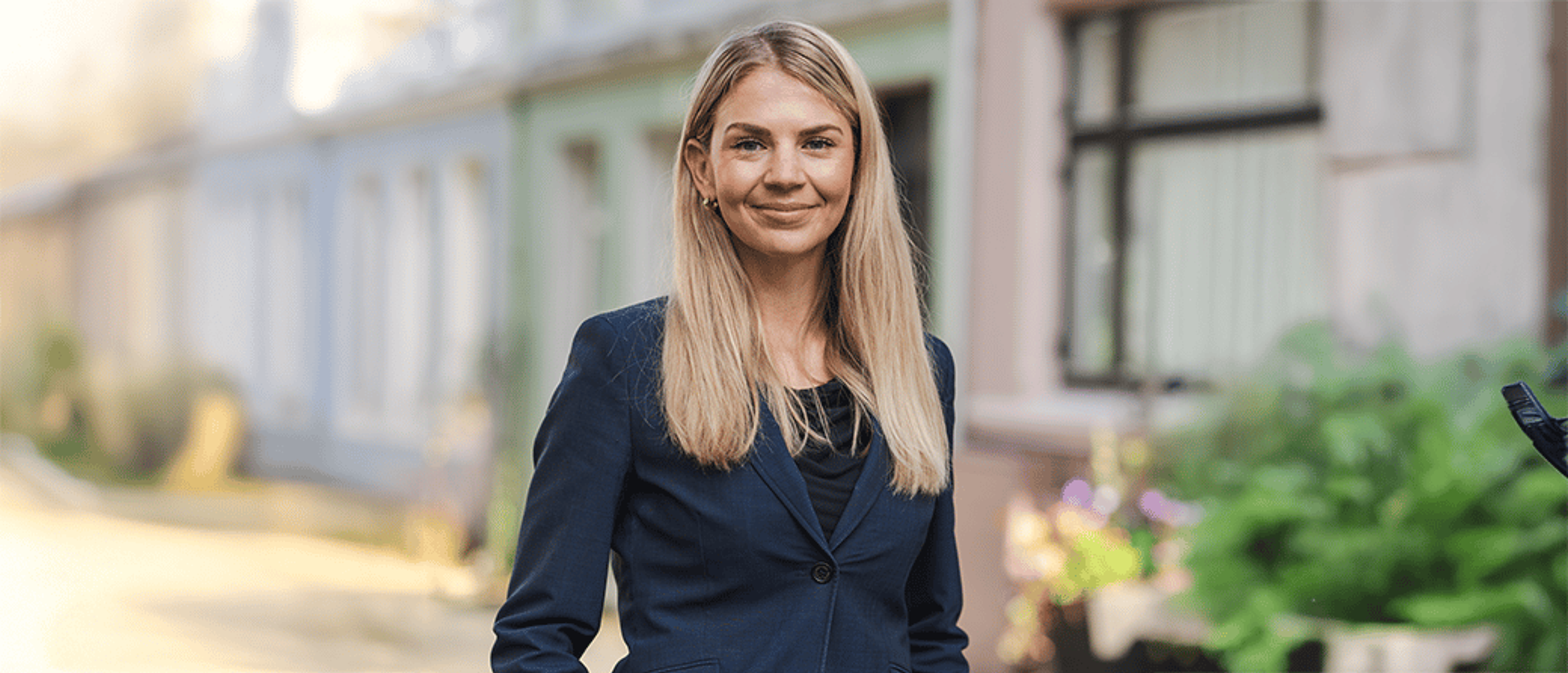 En smilende kvinne med langt, blondt hår og mørk blazer står utendørs foran fargerike bygninger.