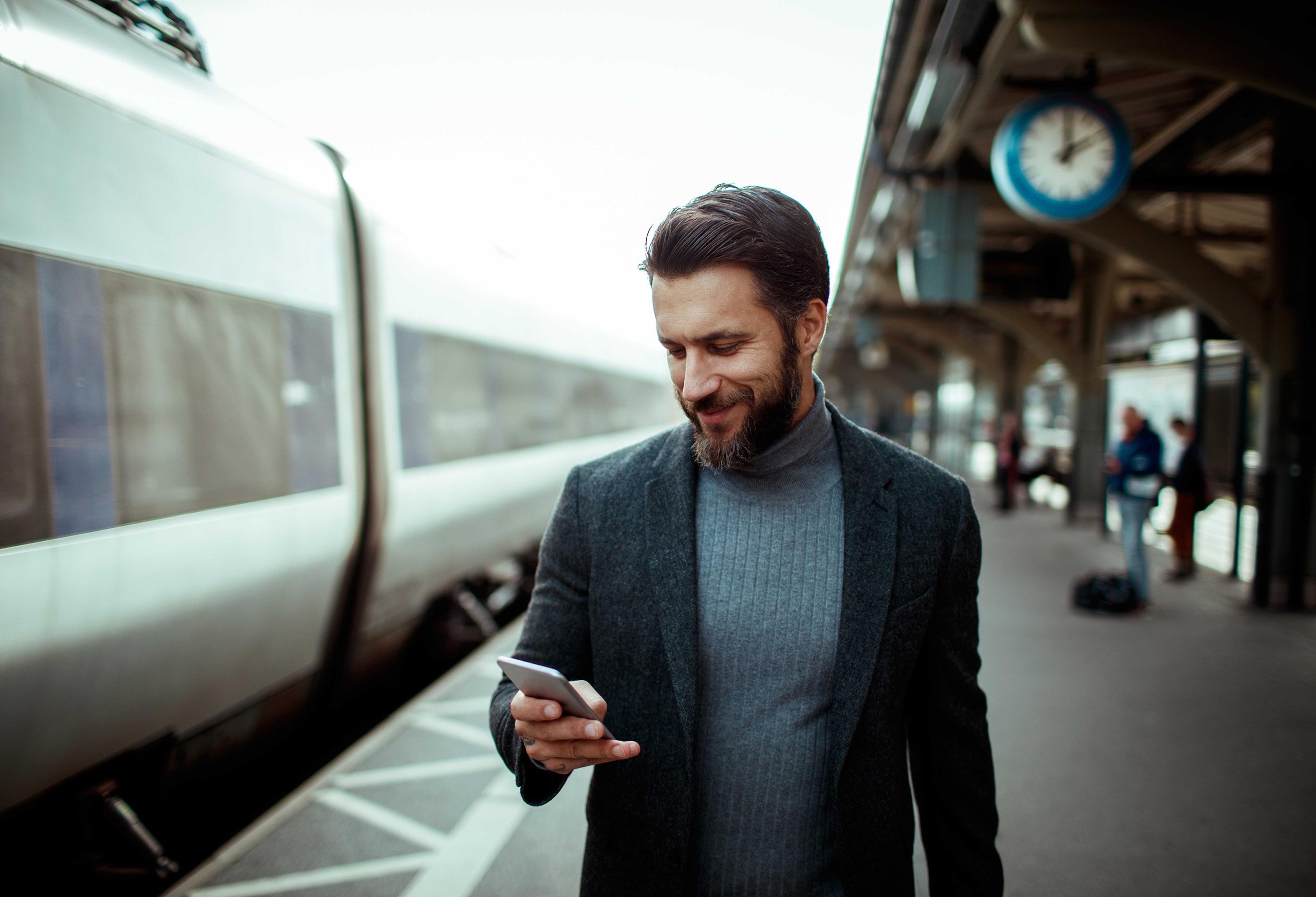 En mann i blazer står på en togperrong ved siden av et tog og ser smilende ned på mobilen sin, mens en stor klokke henger over plattformen i bakgrunnen.