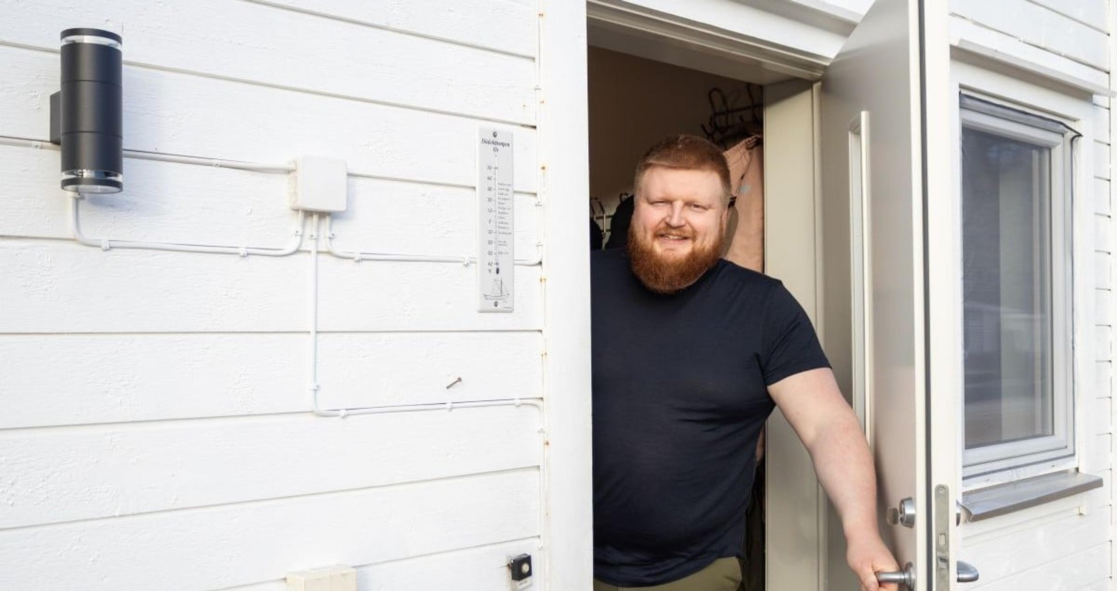 En smilende mann med skjegg står i døråpningen til et hvitt hus og ser ut, med ytterveggens lamper og ledninger synlige ved siden av ham.