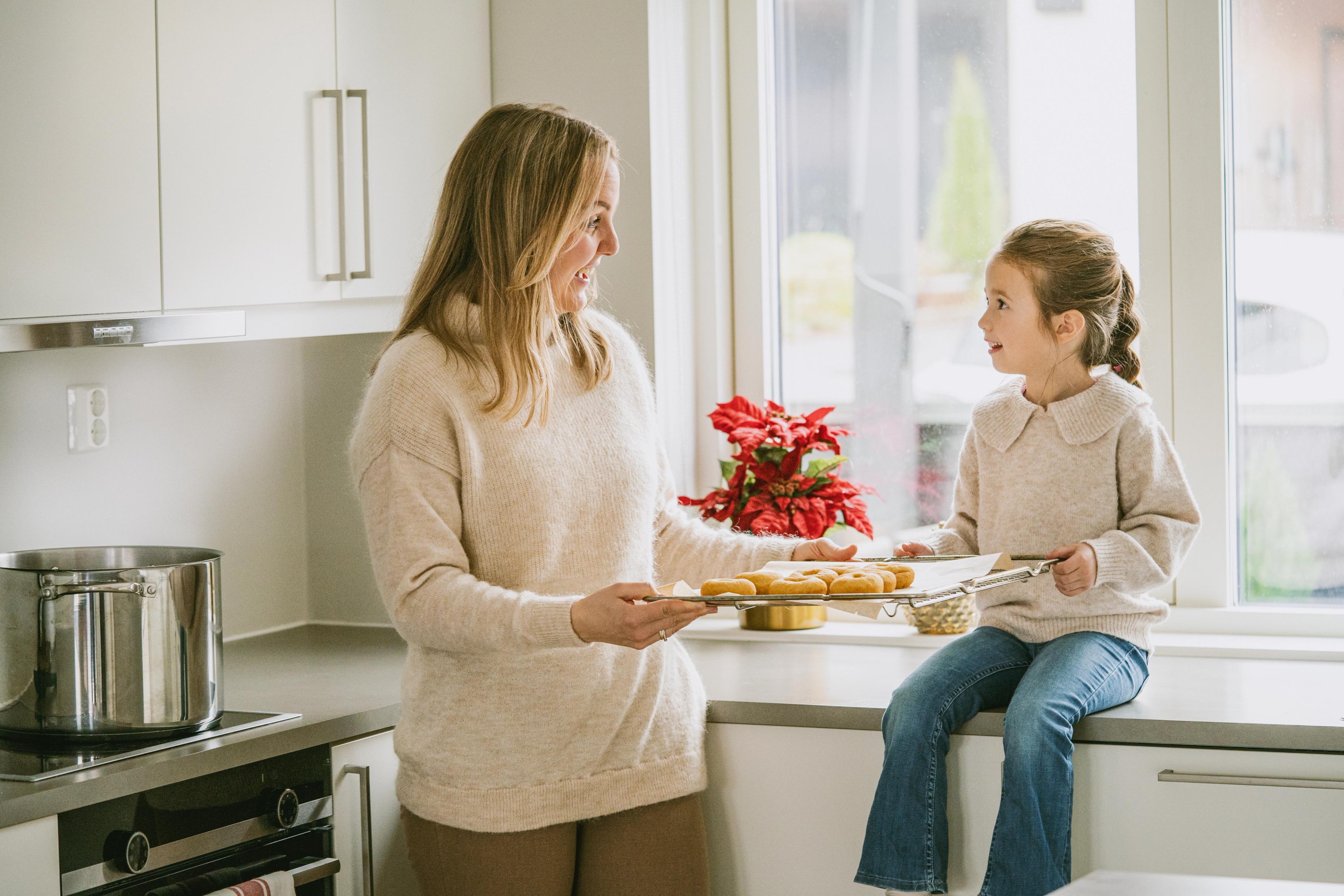 En kvinne og en jente smiler til hverandre mens de holder et stekebrett med nybakte boller på et lyst kjøkken. Jenta sitter på kjøkkenbenken ved vinduet.