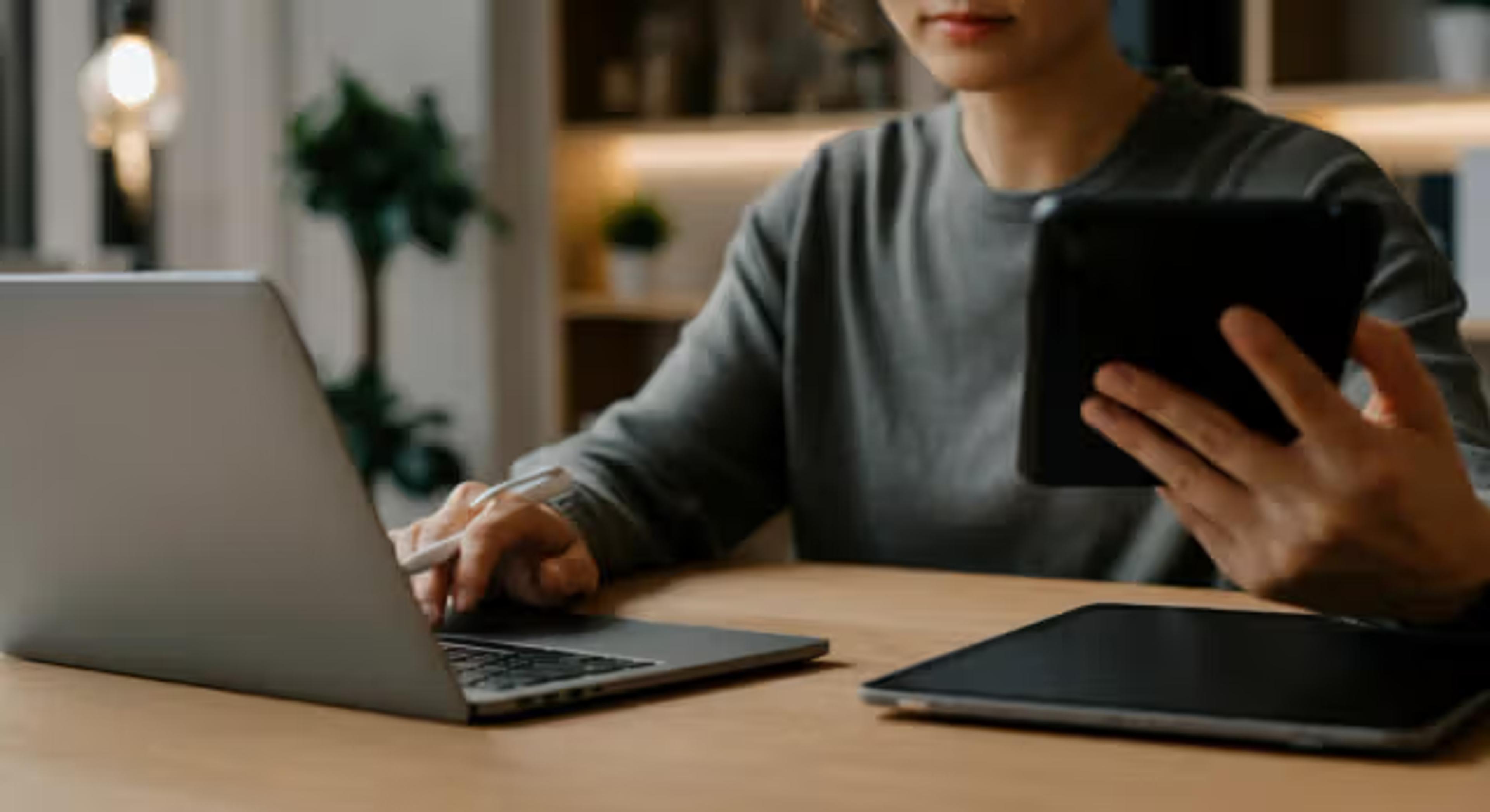 En person sitter ved et bord og jobber på en bærbar PC mens hen holder et nettbrett i den andre hånden, med et annet nettbrett liggende på bordet ved siden av.