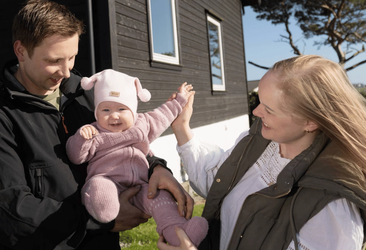 Far som holder baby sammen med mor og alle smiler ute foran et hus i fint vær