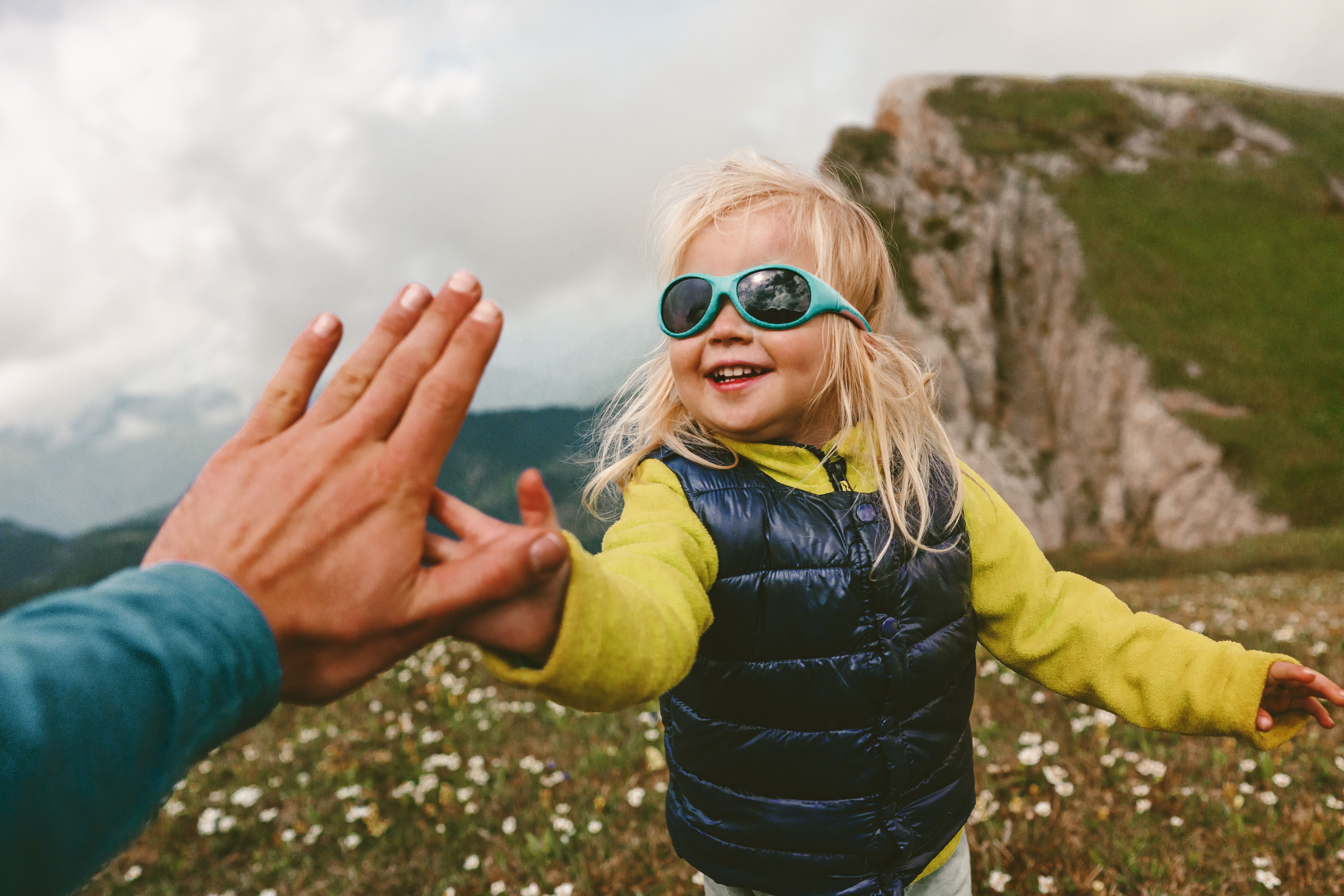 Et smilende, blondt barn med turkise solbriller gir en voksen en high five på en blomstereng i fjellet.