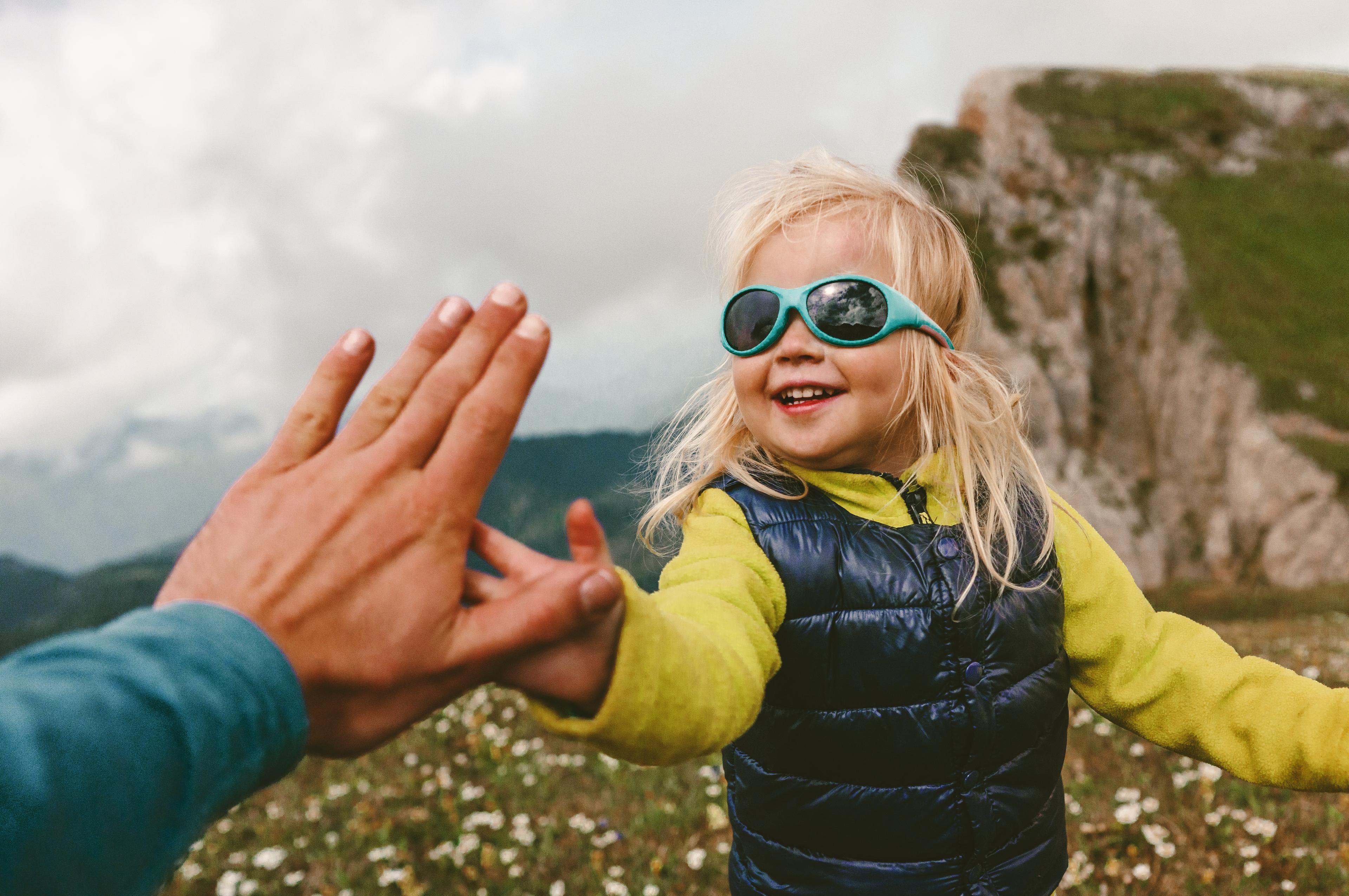 Et smilende, blondt barn med turkise solbriller gir en voksen en high five på en blomstereng i fjellet.