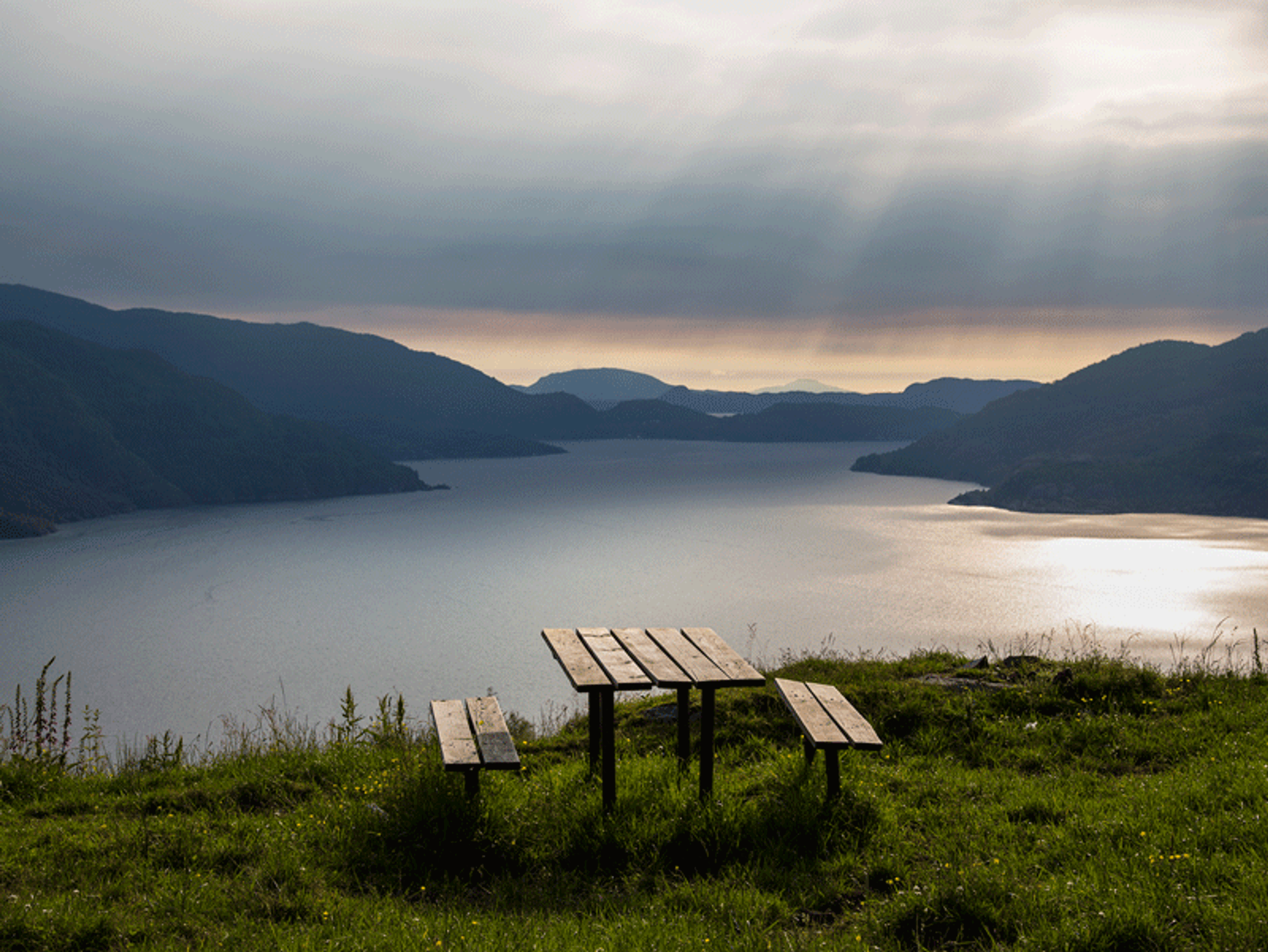 Et enkelt trebord med to benker står på en grønn gresslette med utsikt over en fjord omgitt av mørke fjell under en overskyet himmel.