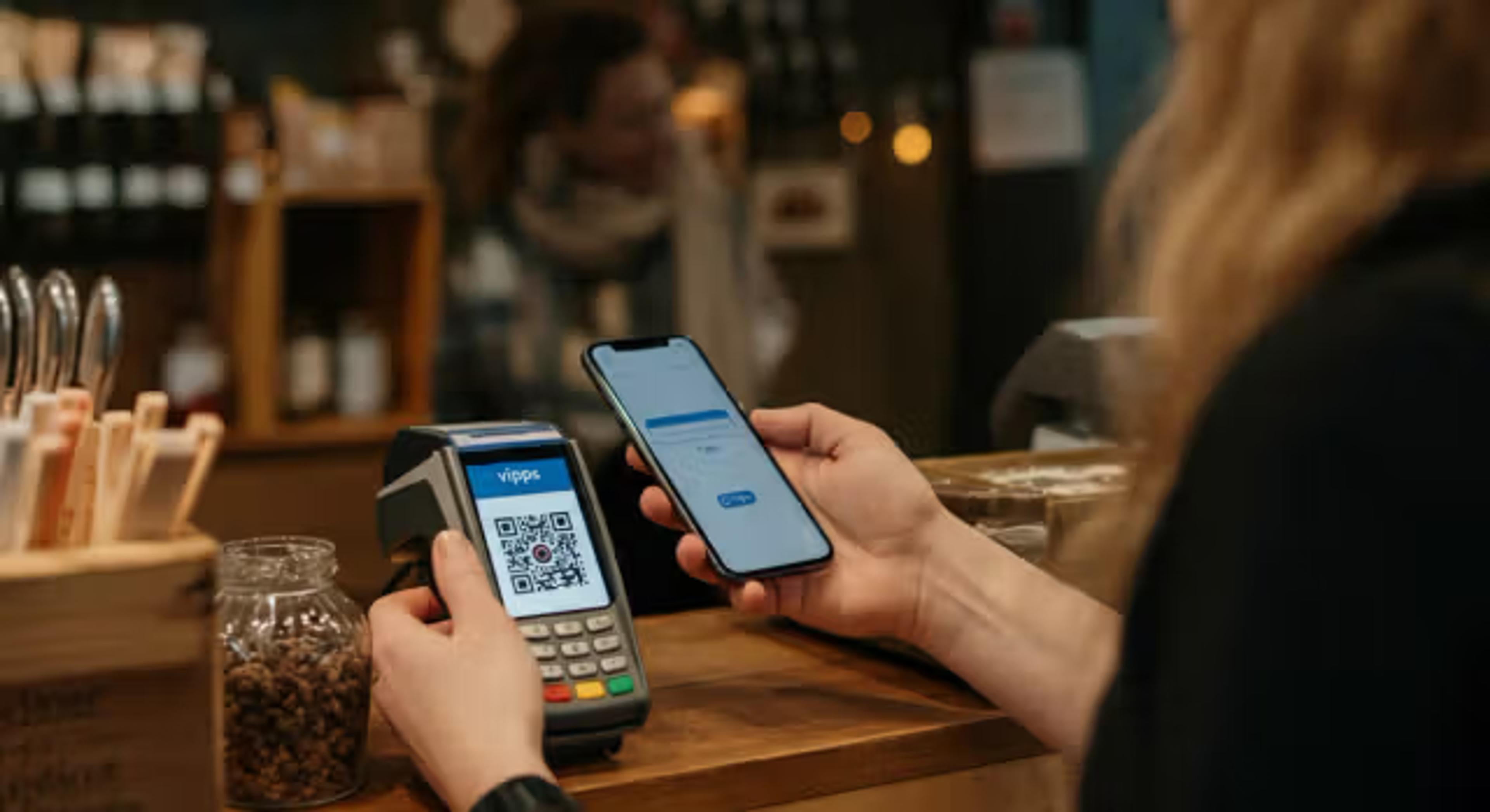 En person står ved en kafédisk og betaler med mobiltelefon ved å skanne en QR-kode på en betalingsterminal merket Vipps.