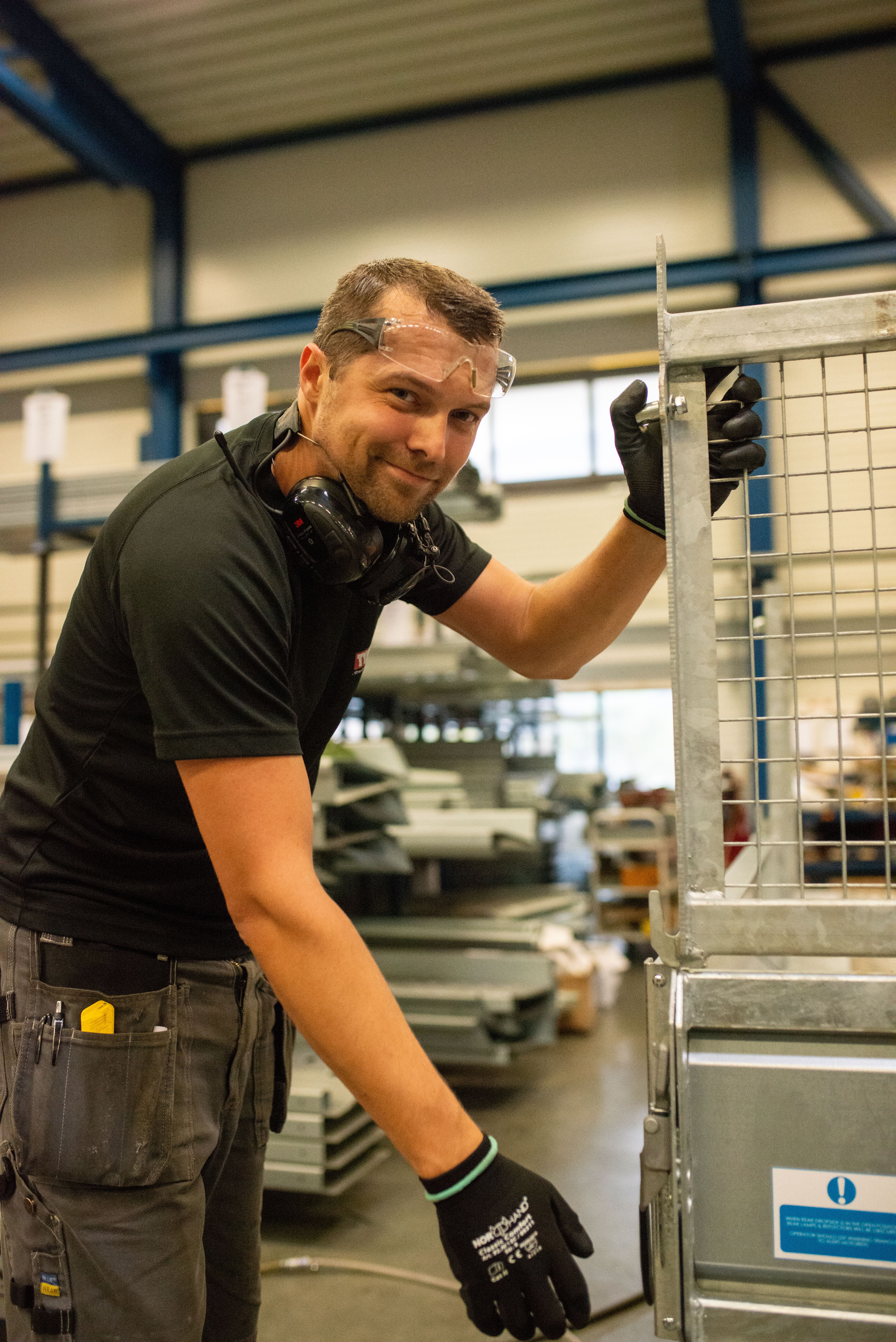 En smilende industriarbeider med vernebriller, hørselsvern og hansker arbeider på en metallbeholder med gitter i en fabrikkhall.
