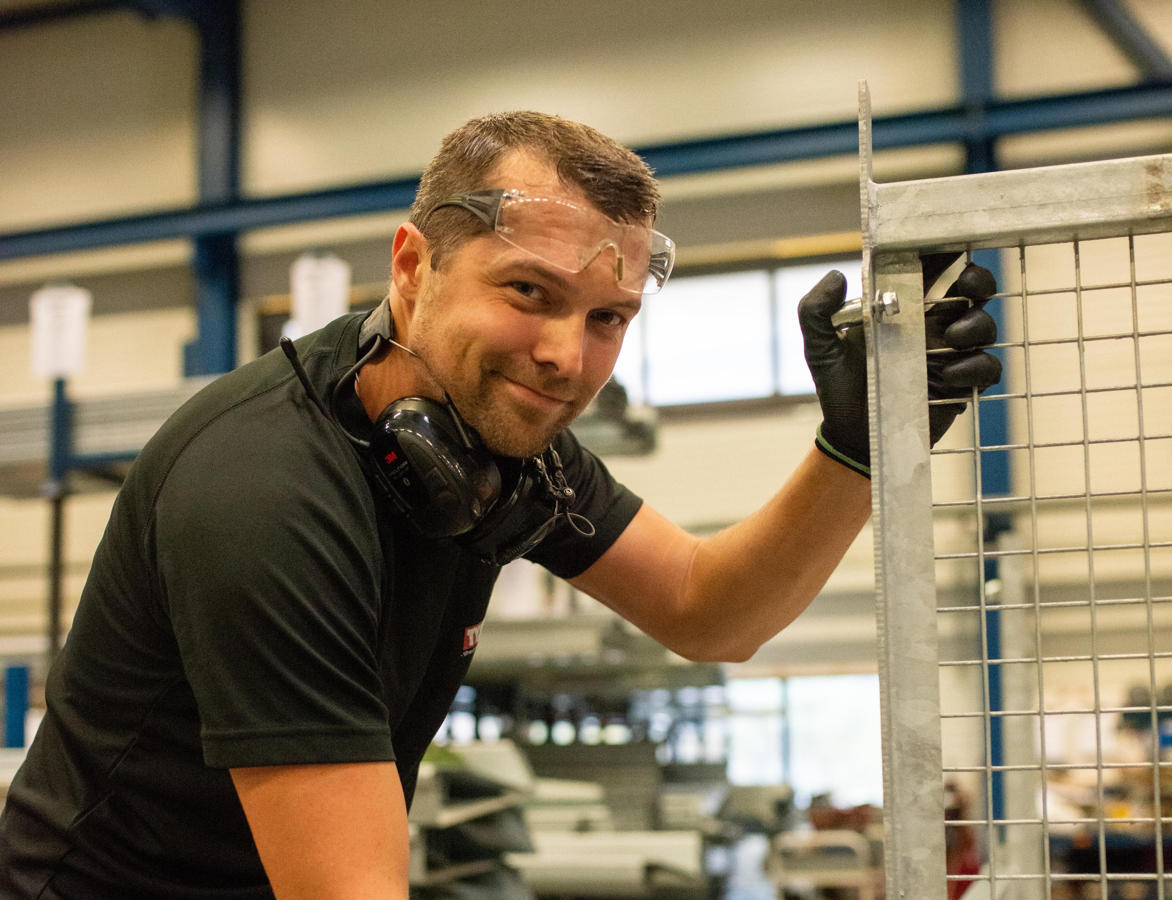 En smilende industriarbeider med vernebriller, hørselsvern og hansker arbeider på en metallbeholder med gitter i en fabrikkhall.