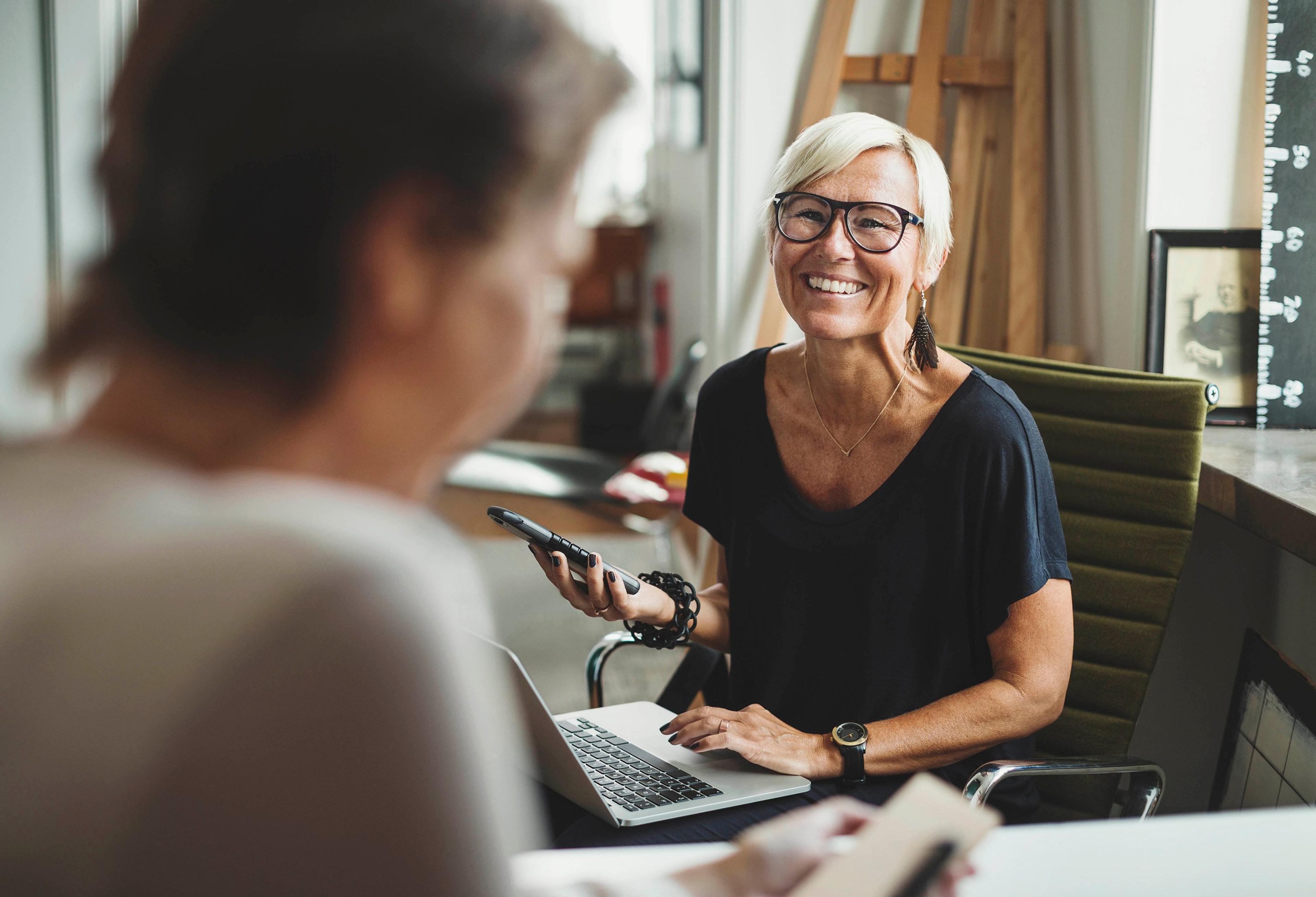 Smilende kvinne med kort, lyst hår og briller sitter ved et skrivebord med en bærbar PC og mobiltelefon, mens hun snakker med en annen person i forgrunnen.