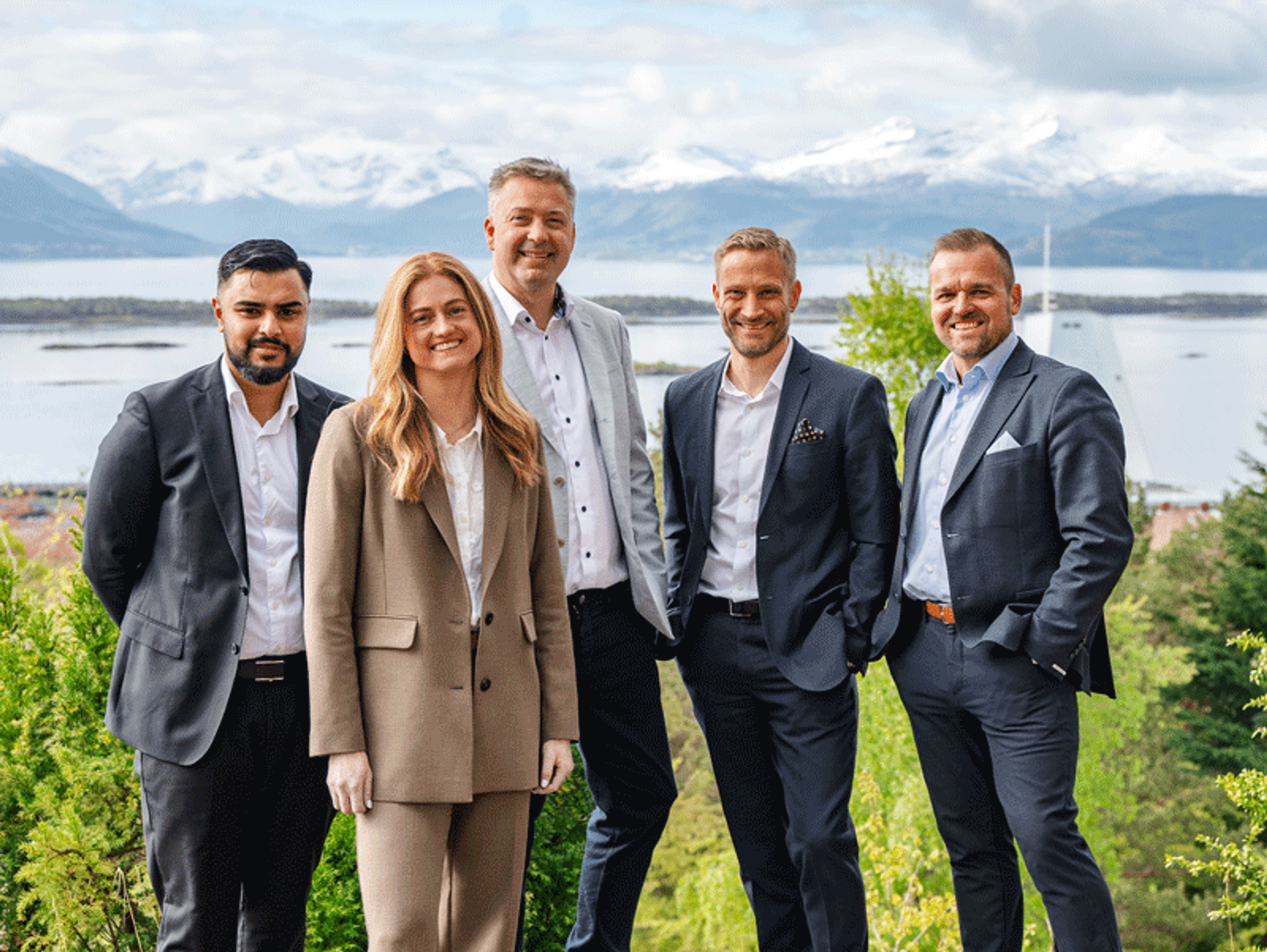 Fem forretningskledde personer står sammen utendørs og smiler mot kameraet, med fjord og snødekte fjell i bakgrunnen.
