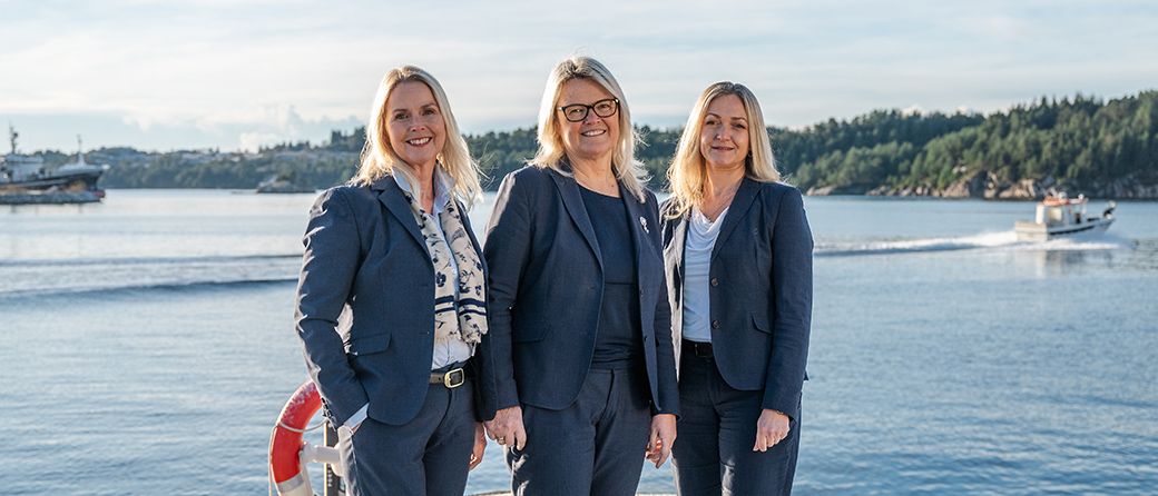 Tre smilende kvinner i mørkeblå dresser står ved sjøen, med skogkledde øyer og en liten båt i bakgrunnen.
