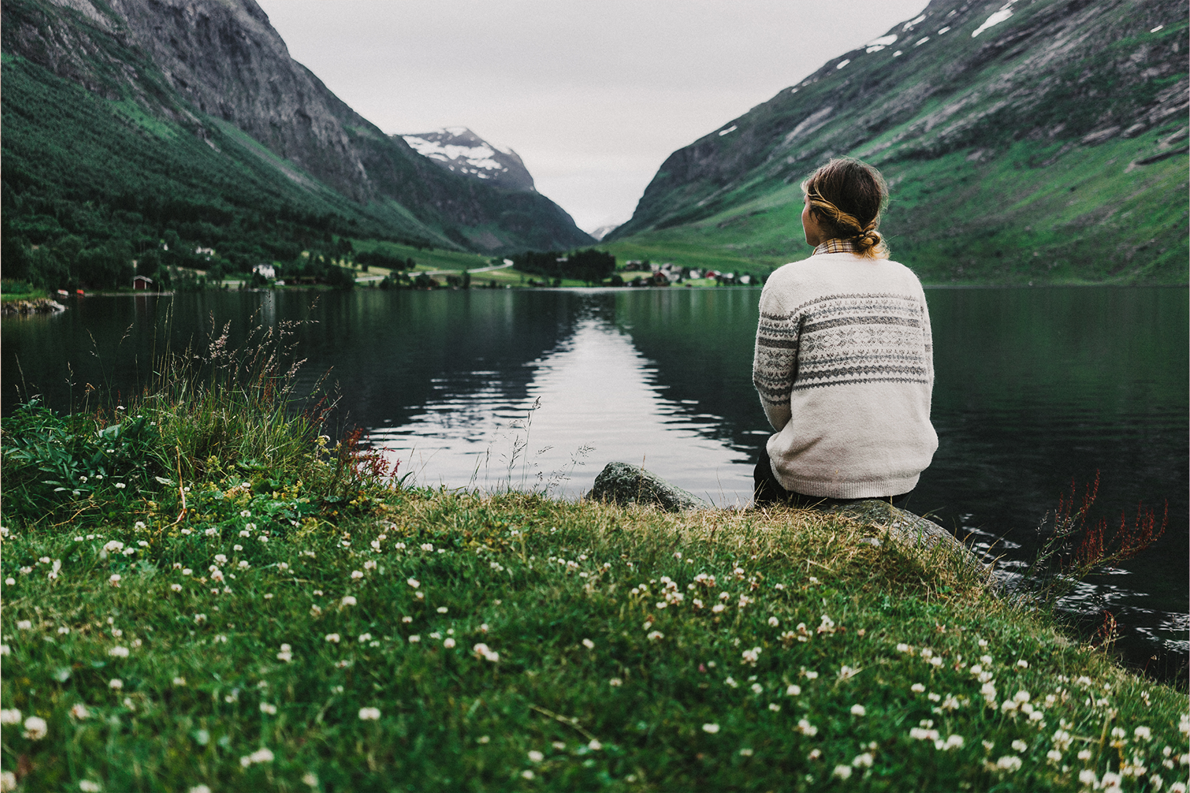 En person i mønstret ullgenser sitter ved bredden av en stille fjord og ser mot grønne, bratte fjell med snøflekker.