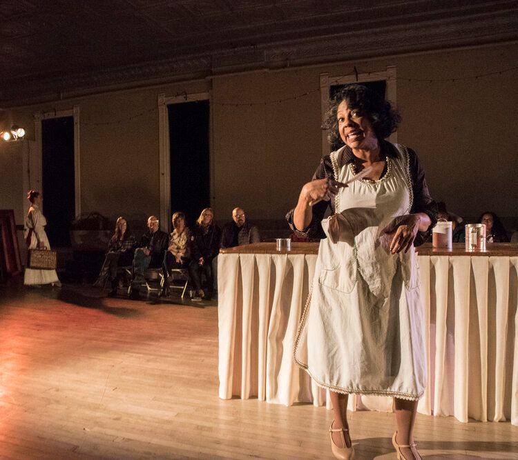 a Black woman in an apron, singing into a wooden spoon